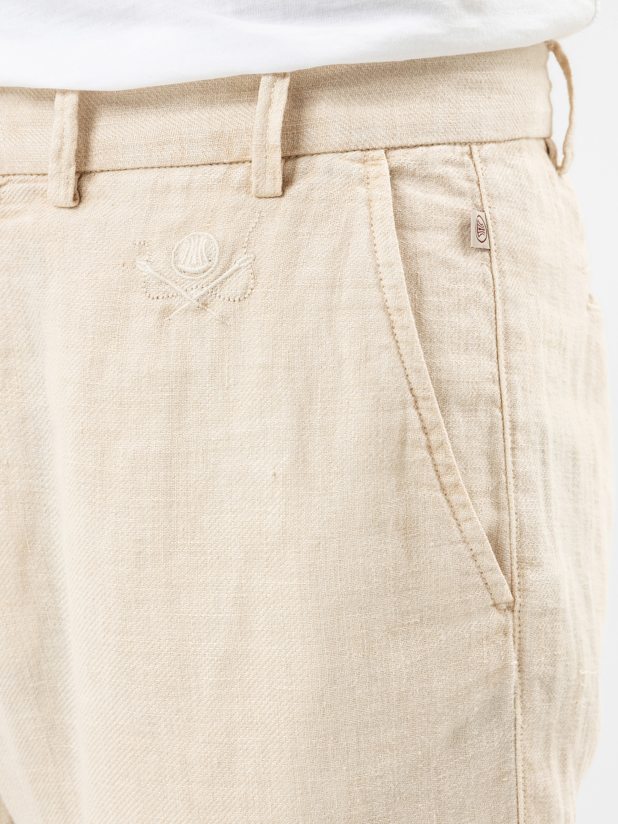 Nahaufnahme einer Person, die eine hellbeige Leinenhose mit sichtbarer Seitentasche, Gürtelschlaufen und einem dezenten gestickten Logo in der Nähe des Hosenbundes trägt, gepaart mit einem weißen Hemd, das in die Hose gesteckt wird.