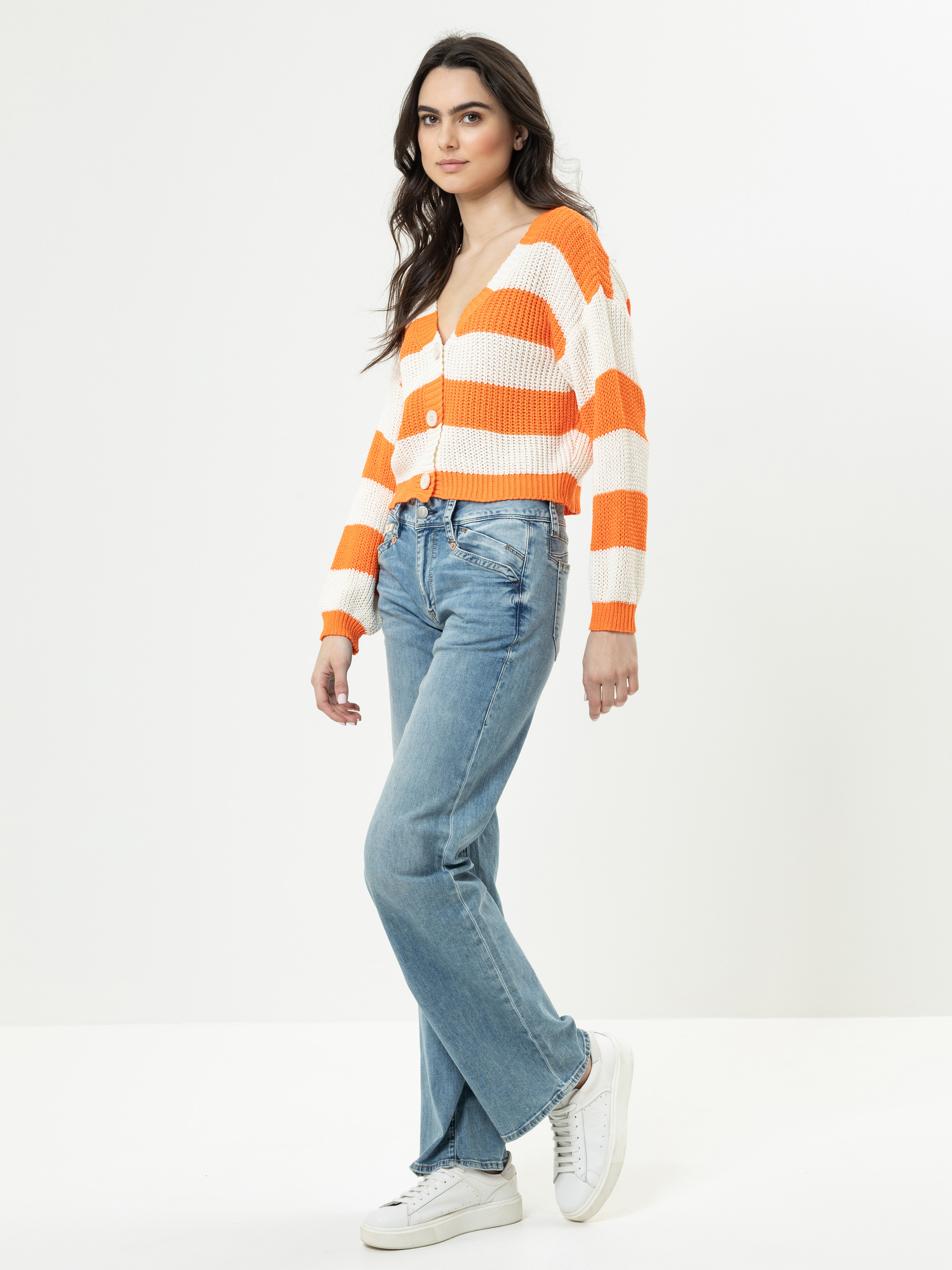 Eine Frau steht vor einem schlichten weißen Hintergrund und trägt eine orange-weiß gestreifte Strickjacke, blaue Jeans und weiße Turnschuhe. Sie hat langes, gewelltes dunkles Haar und einen entspannten Gesichtsausdruck.