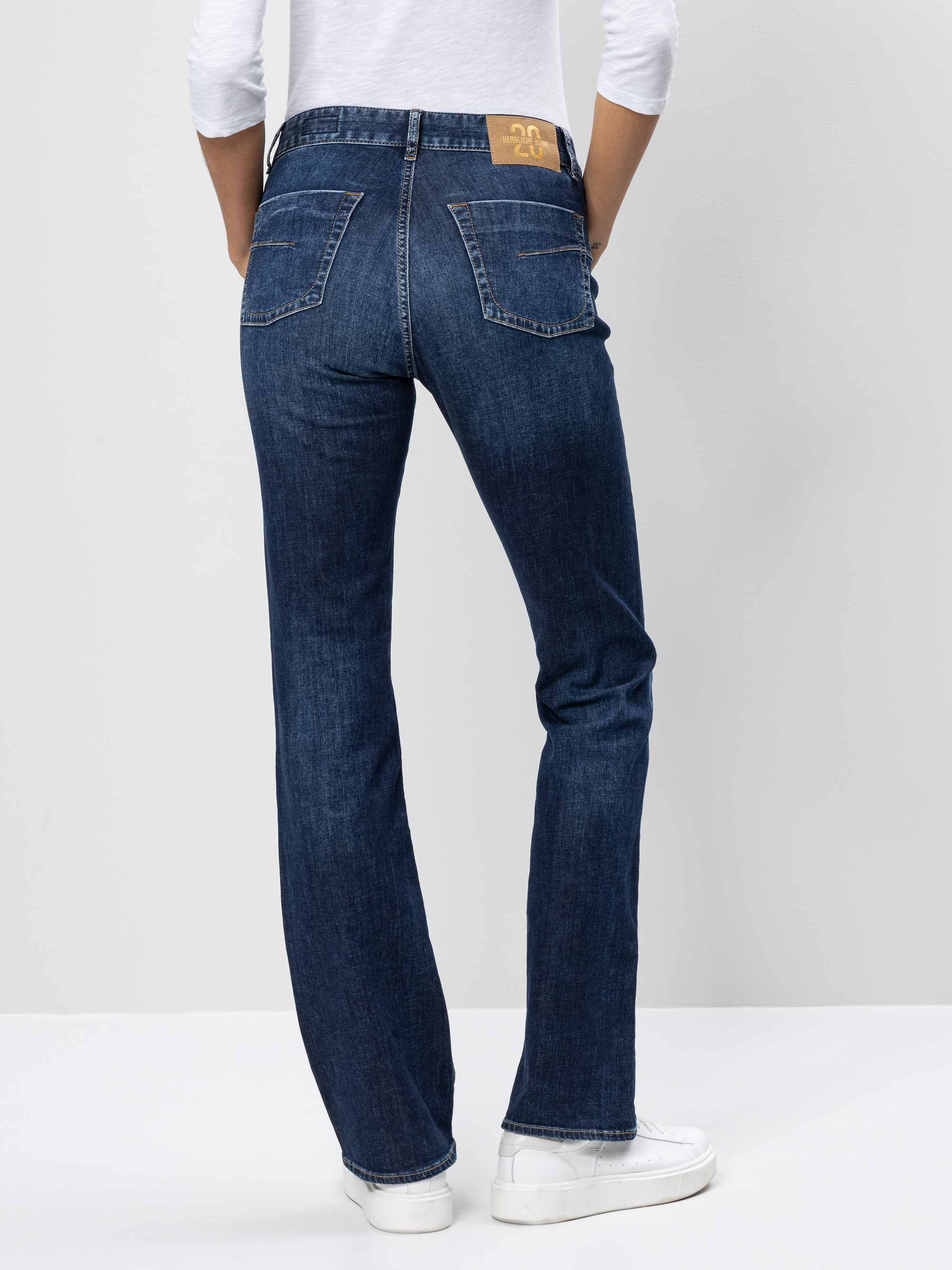 Eine Person, die eine dunkelblaue Jeans mit geradem Bein, ein weißes langärmeliges Hemd und weiße Turnschuhe trägt, steht mit dem Gesicht nach hinten, die Hände in die Gesäßtaschen gestützt, vor einem schlichten hellen Hintergrund.