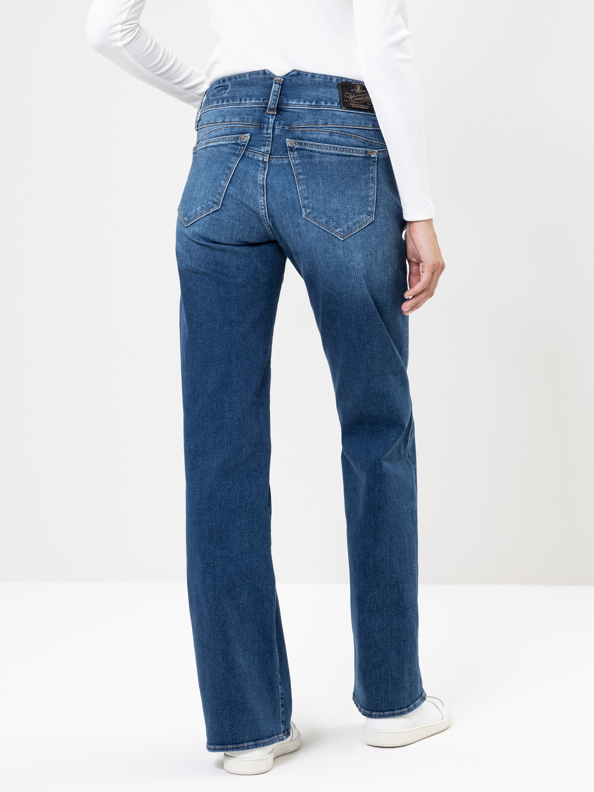 Eine Person, die eine blaue Jeans mit geradem Bein, ein weißes langärmeliges Oberteil und weiße Turnschuhe trägt, steht mit dem Rücken zur Kamera vor einem schlichten hellen Hintergrund.