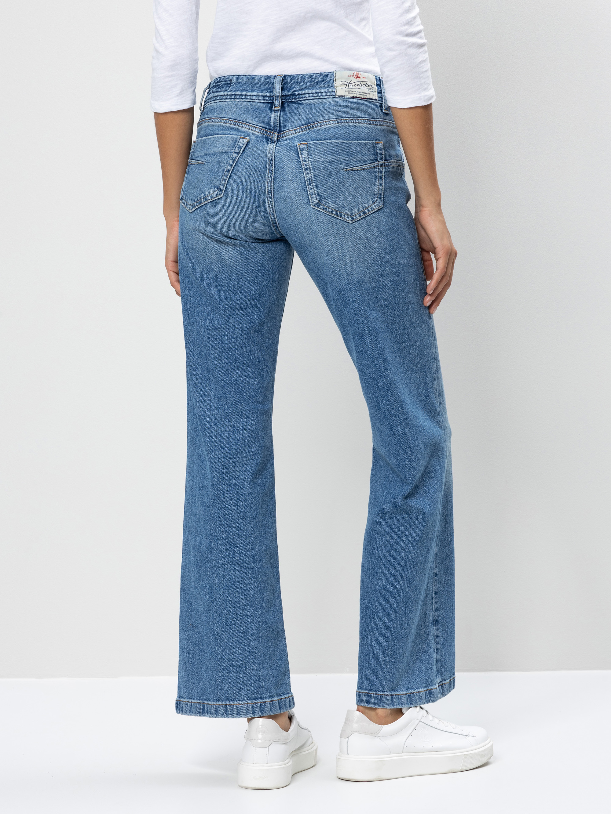 Eine Person, die eine blaue Jeans mit geradem Bein, ein weißes langärmeliges Hemd und weiße Turnschuhe trägt, steht mit dem Gesicht nach hinten und zeigt die Rückenansicht der Jeans vor einem neutralen Hintergrund.