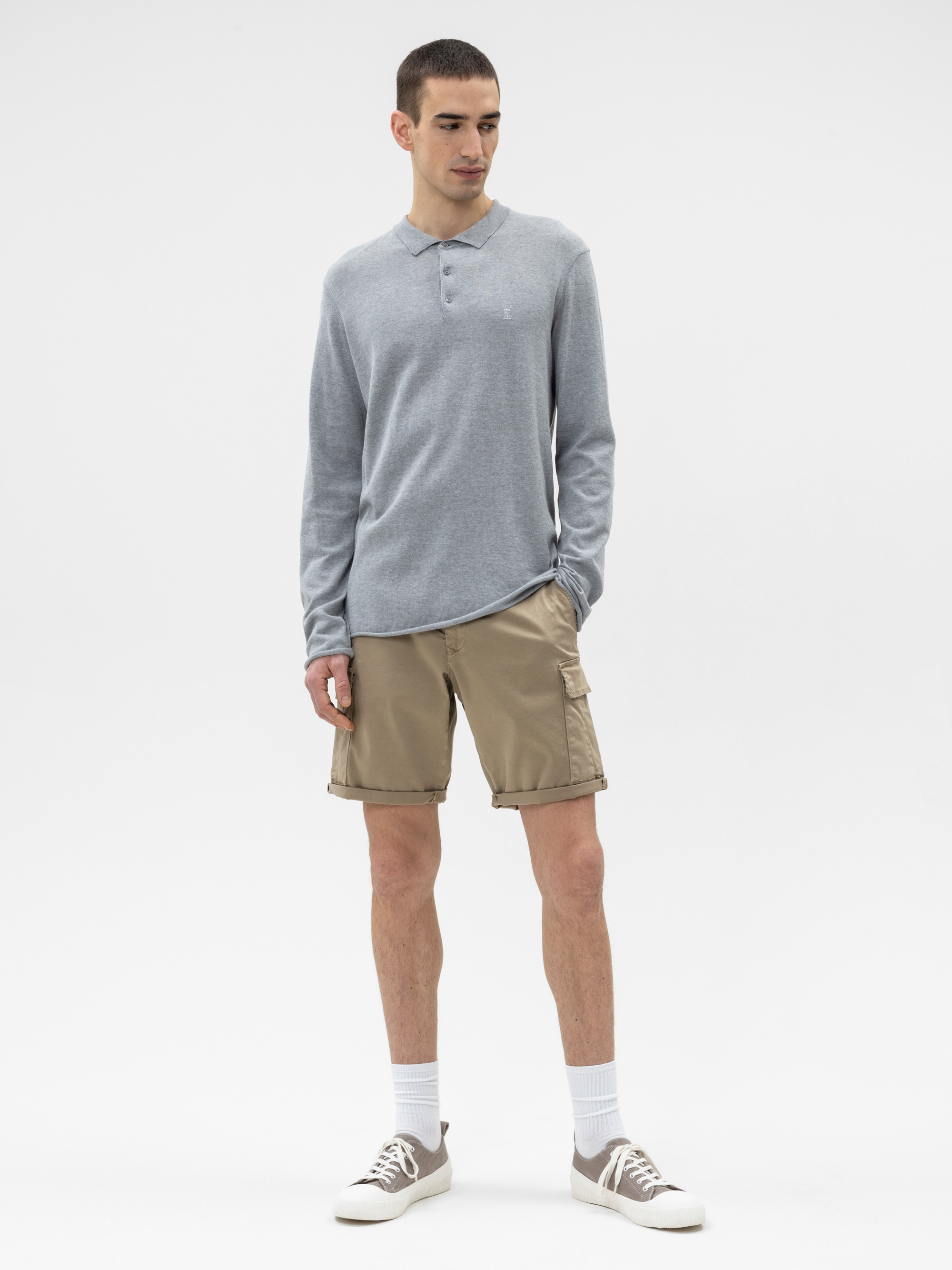 Ein Mann steht vor einem weißen Hintergrund und trägt ein hellgraues, langärmeliges Poloshirt, khakifarbene Cargo-Shorts, weiße Socken und graue Turnschuhe. Er hat kurze Haare und blickt leicht nach links.