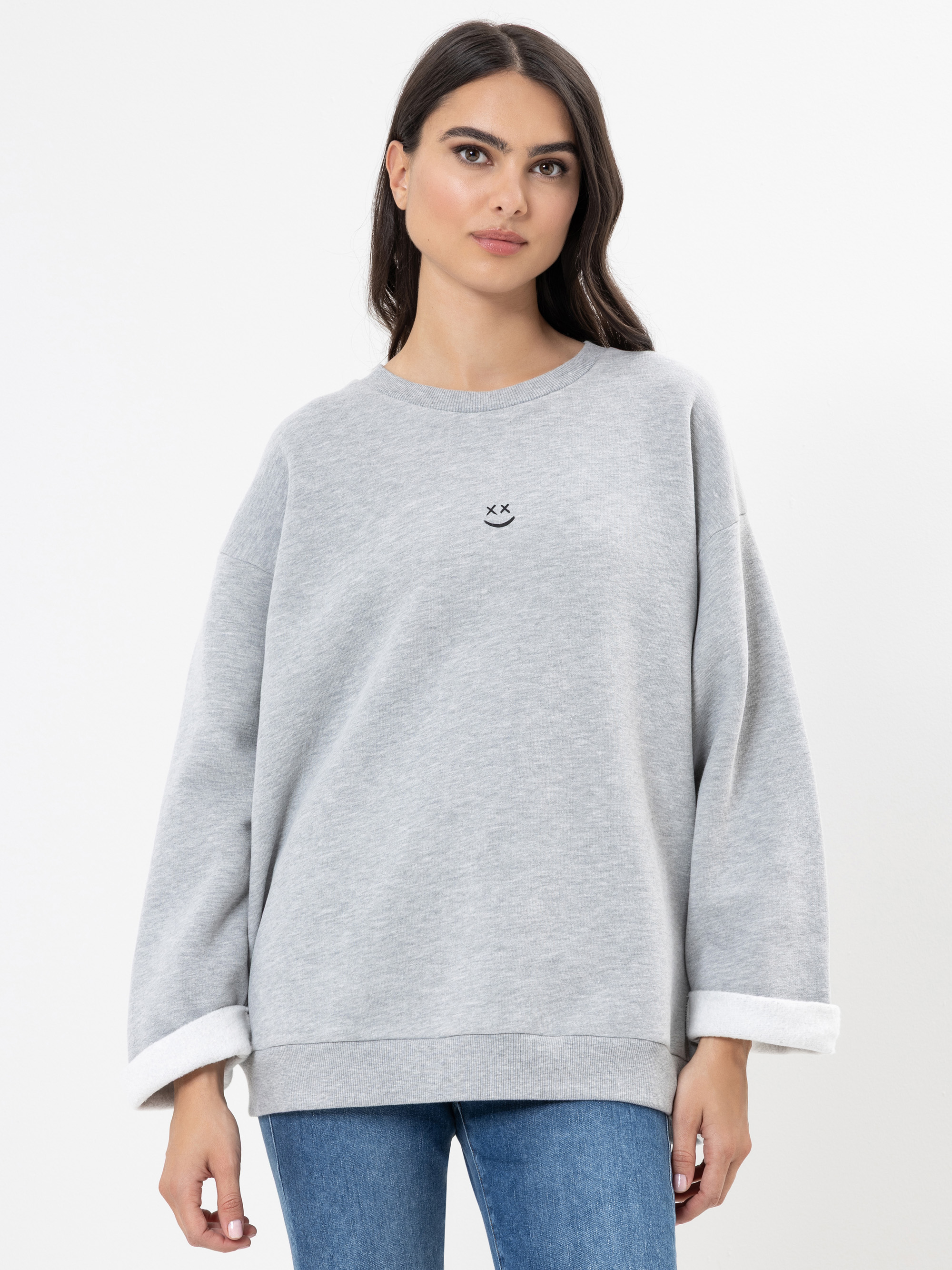 Eine Frau mit langen dunklen Haaren trägt ein locker sitzendes, hellgraues Sweatshirt mit einem kleinen, einfachen Gesichtsmuster auf der Brust. Sie steht vor einem schlichten weißen Hintergrund und schaut in die Kamera.