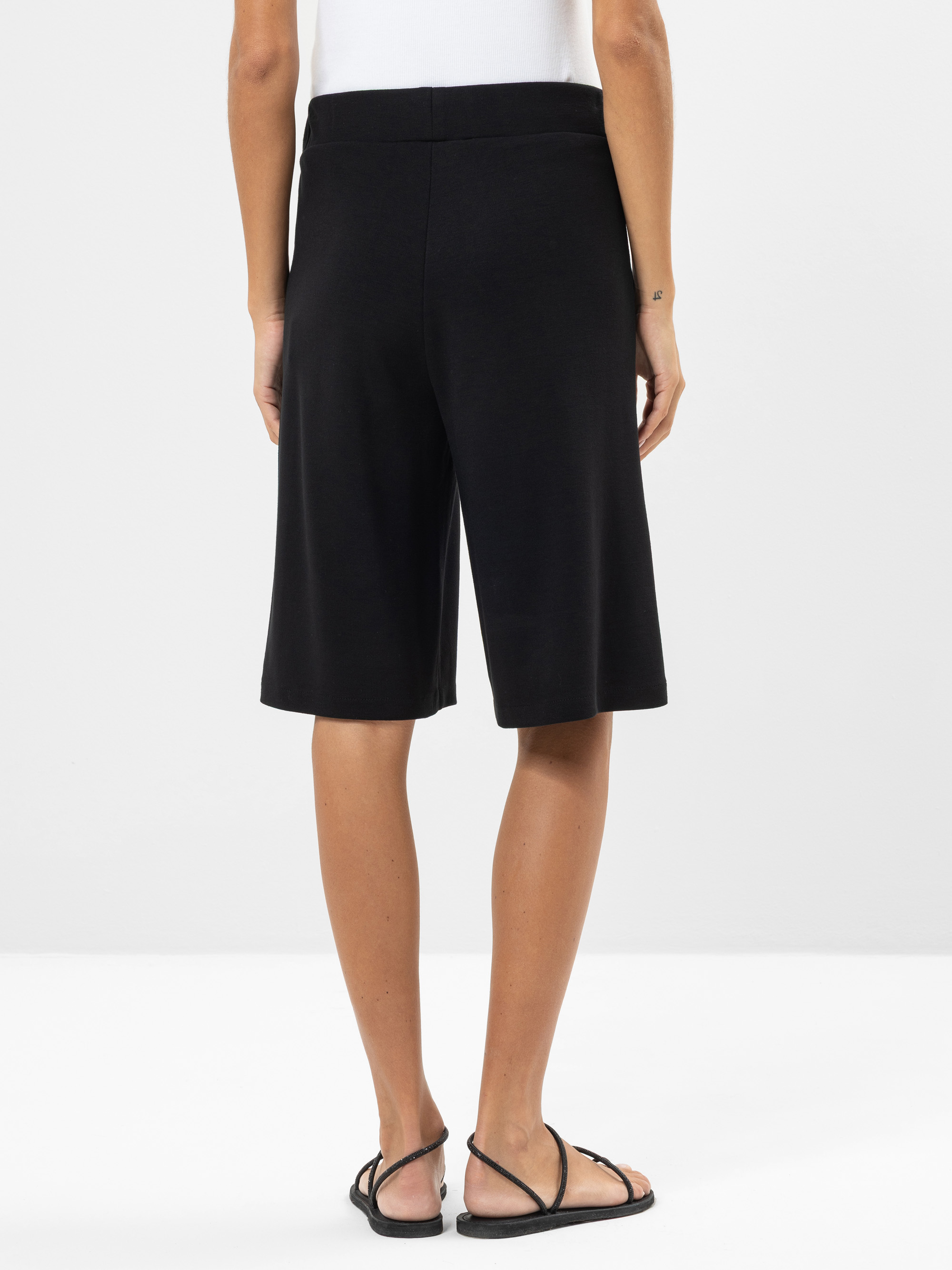 Eine Person, die schwarze knielange Shorts, ein weißes Oberteil und schwarze Sandalen trägt, steht mit dem Gesicht zur Kamera vor einem schlichten weißen Hintergrund.