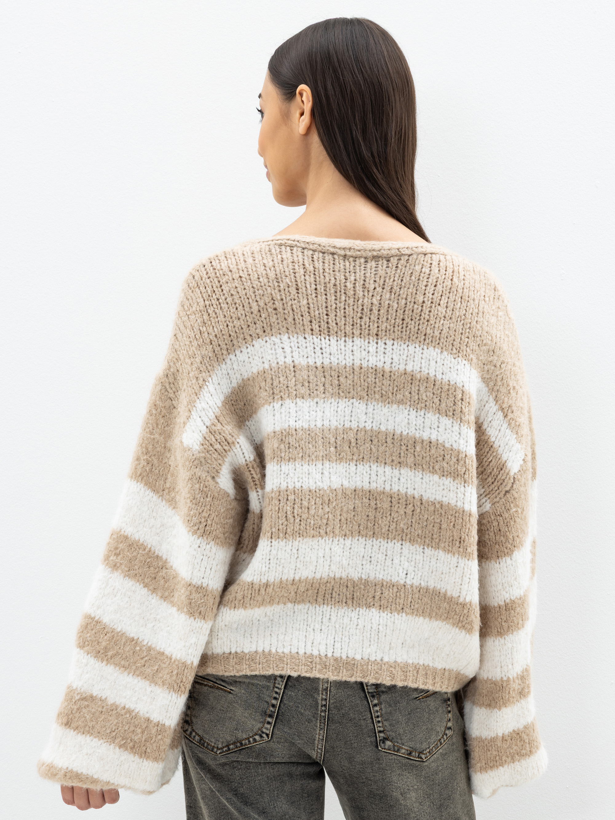 Eine Frau mit langen dunklen Haaren, von hinten gesehen, trägt einen lockeren Strickpulli mit breiten beigen und weißen Querstreifen und ein Paar verblichene graue Jeans, die vor einem schlichten weißen Hintergrund stehen.