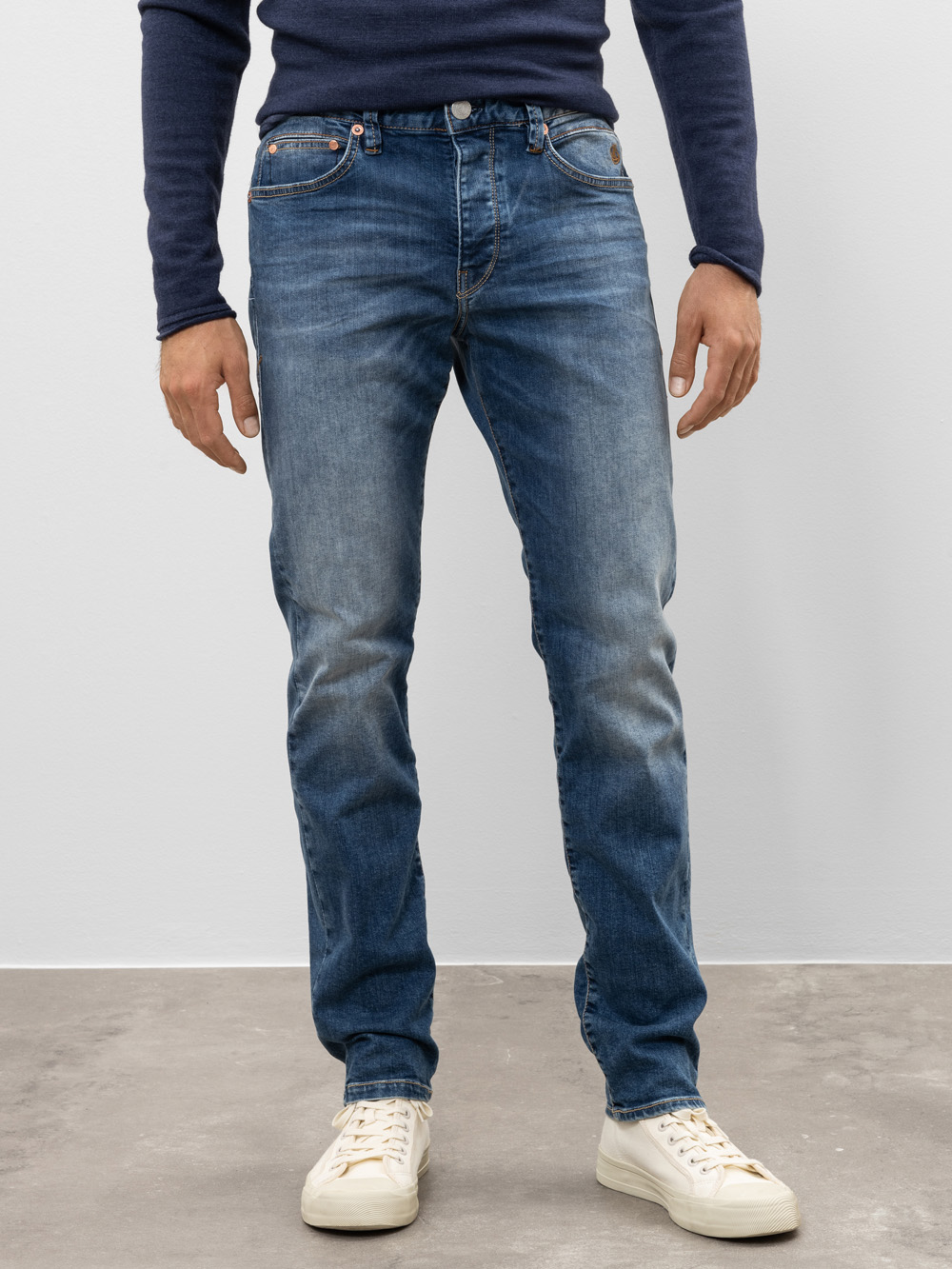 Eine Person, die ein marineblaues langärmeliges Hemd, blaue Jeans und weiße Turnschuhe trägt, steht auf einem grauen Boden vor einer schlichten weißen Wand. Das Foto ist von den Schultern abwärts beschnitten.