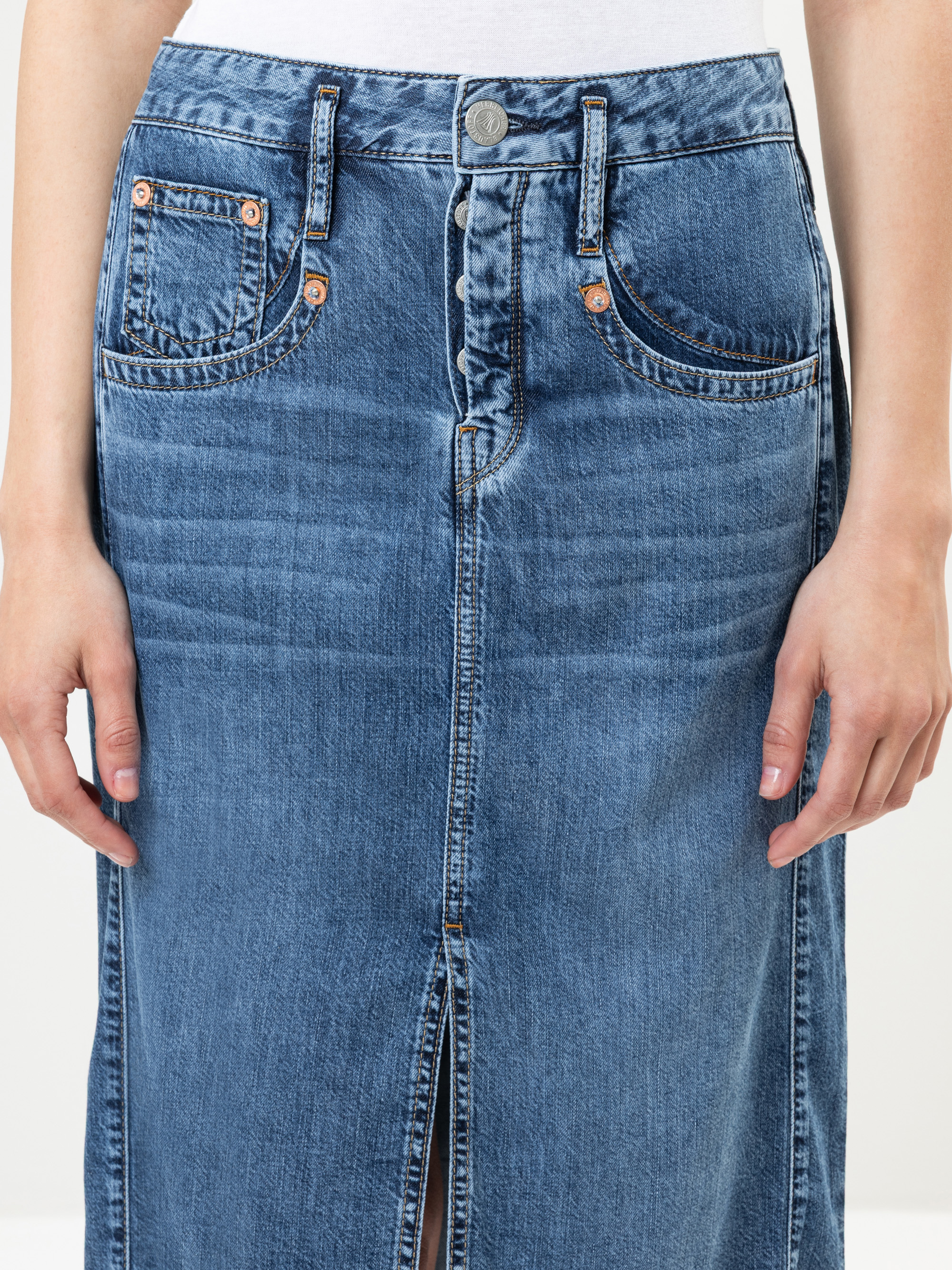 Eine Person trägt einen blauen Jeansrock mit Vordertaschen, Kupfernieten, Gürtelschlaufen, Knopfverschluss und einem Schlitz vorne. Die Hände der Person liegen entspannt an der Seite.