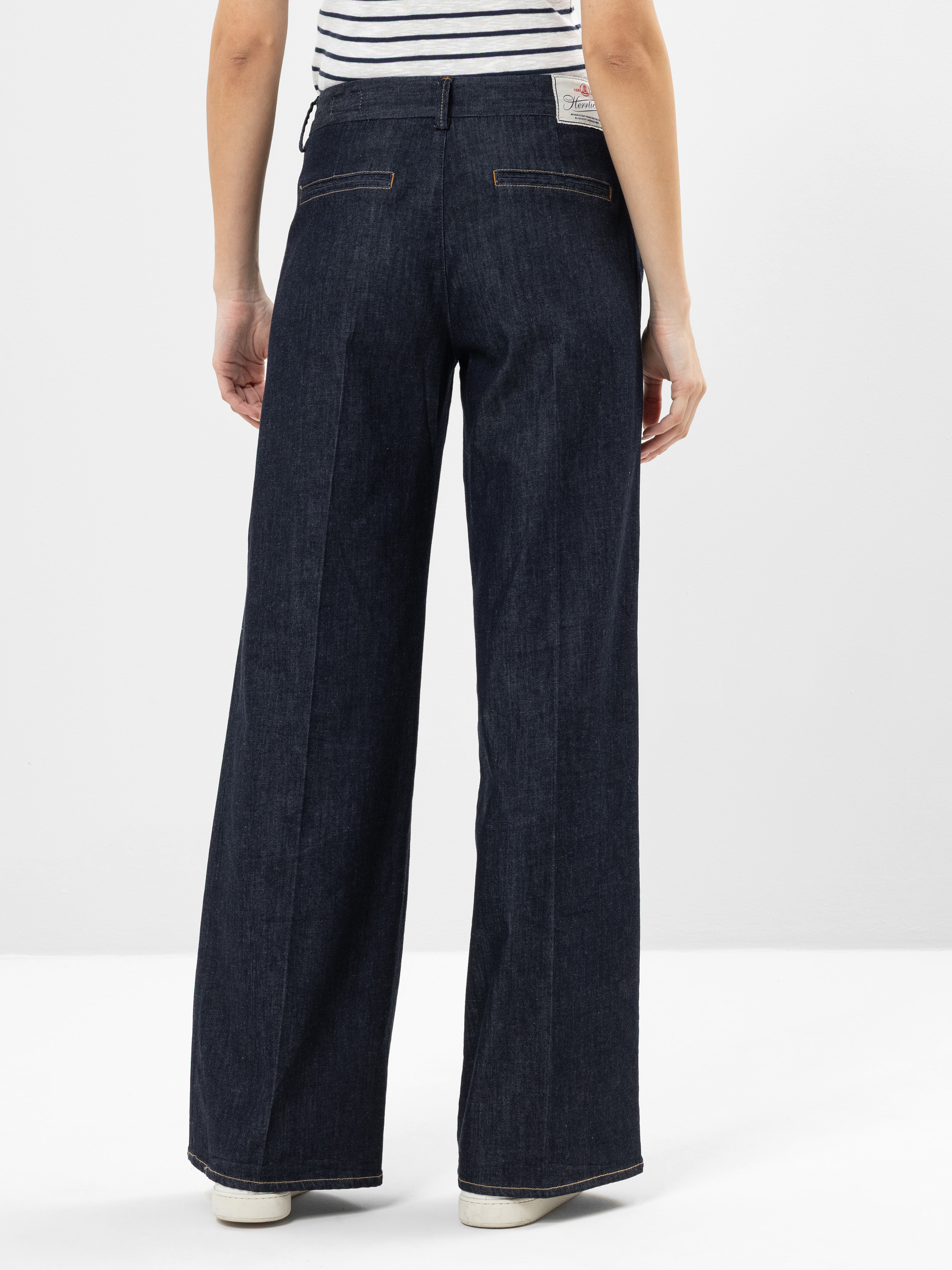 Eine Person steht mit dem Gesicht nach hinten und trägt eine dunkle Jeans mit weitem Bein und sichtbaren Nähten sowie ein gestreiftes kurzärmeliges Hemd. Die Jeans hat Leistentaschen auf der Rückseite und ein weißes Etikett am Bund. Der Hintergrund i