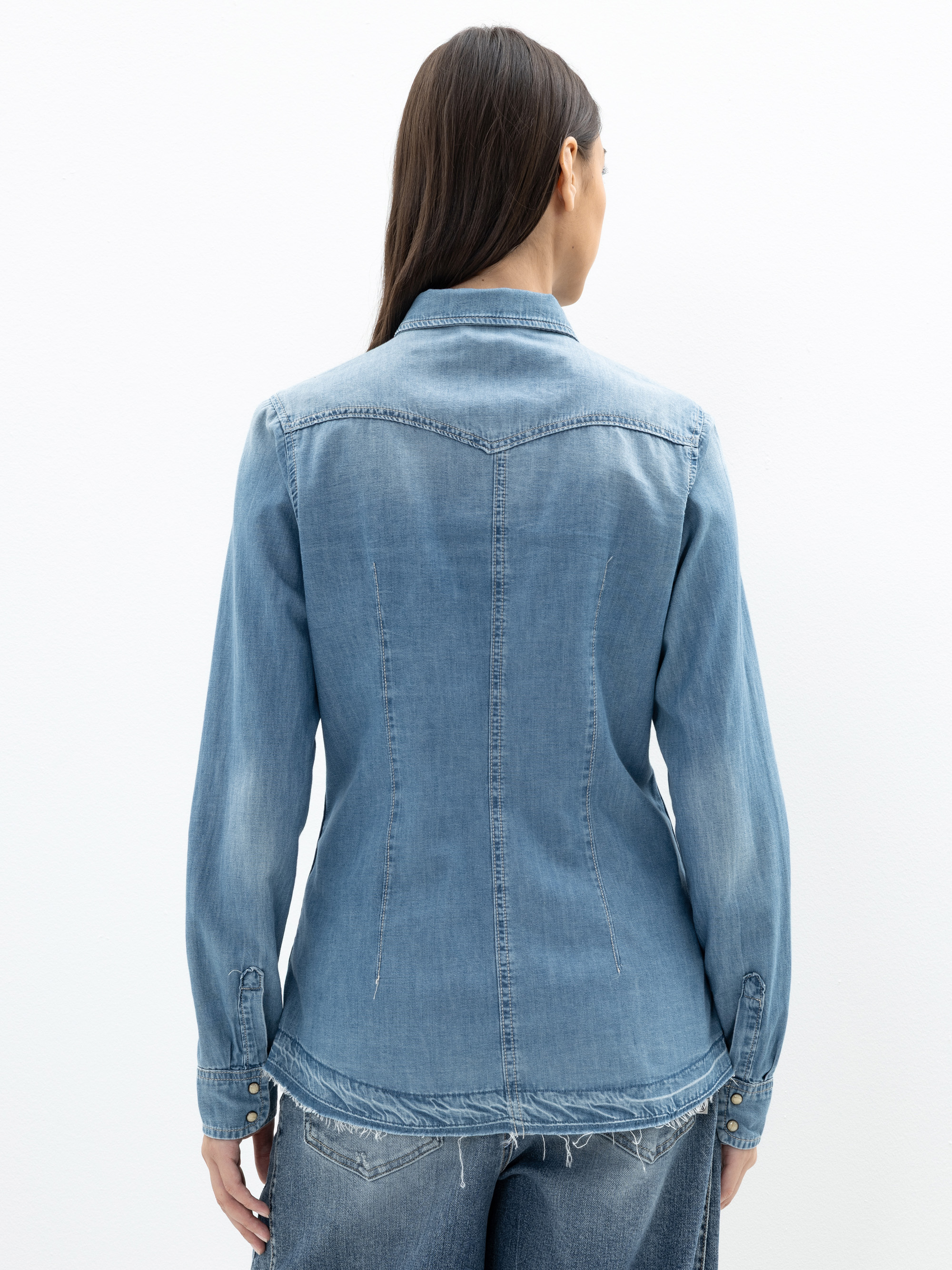 Eine Frau mit langen braunen Haaren trägt ein hellblaues Jeanshemd und blaue Jeans und steht mit dem Rücken zur Kamera vor einem schlichten weißen Hintergrund.