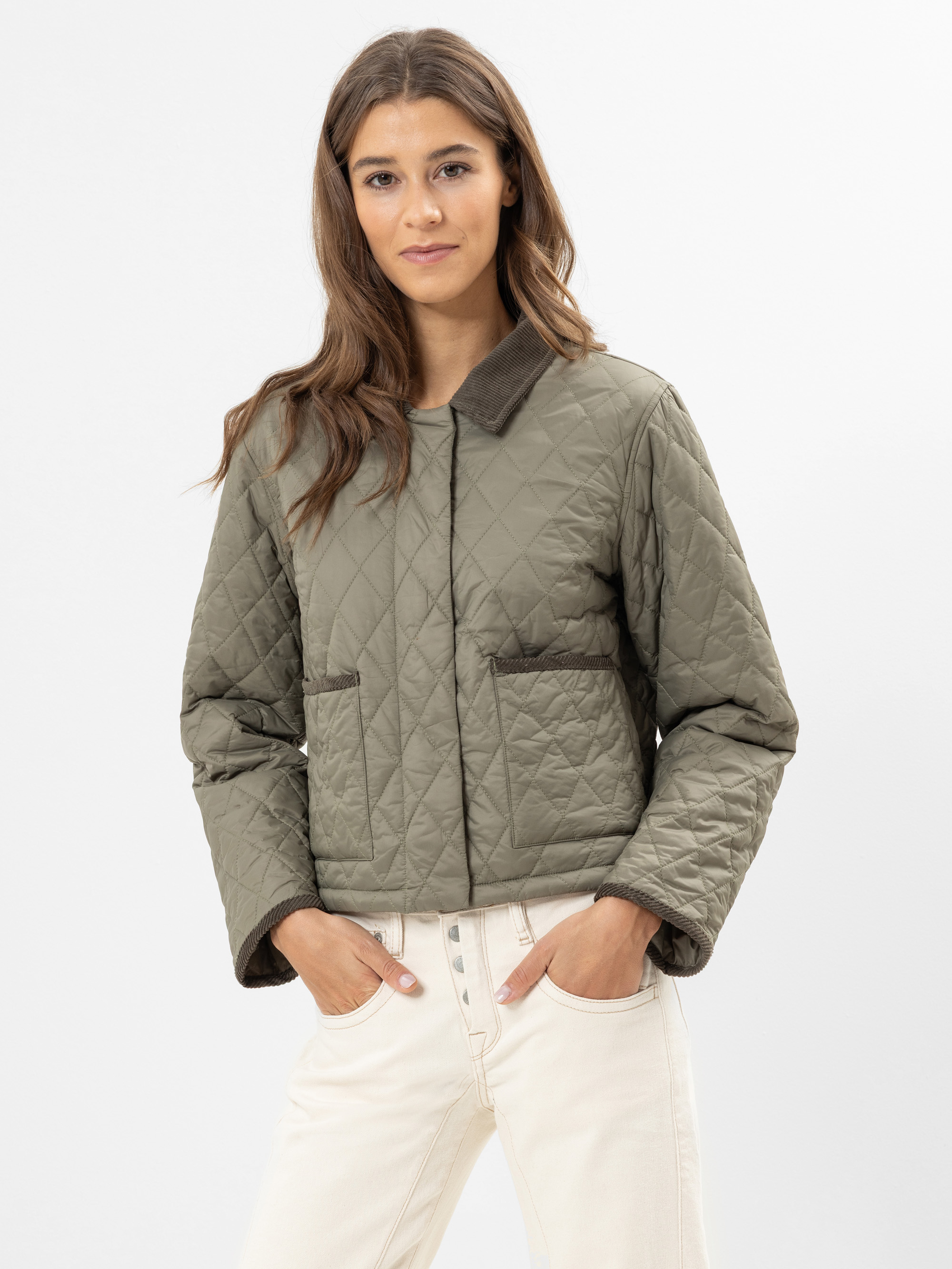 Eine Frau in einer gesteppten, olivgrünen Jacke mit braunem Kragen und Saum steht vor einem schlichten weißen Hintergrund. Sie hat langes braunes Haar und ist mit einer hellen Hose bekleidet.