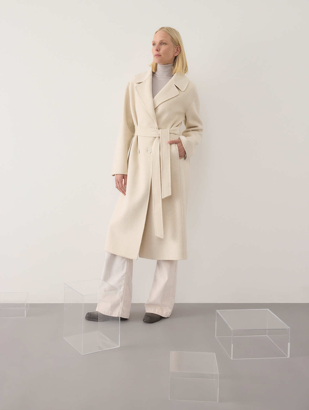 Eine Frau, die einen langen, cremefarbenen Mantel mit Gürtel über einem hellen Rollkragenpullover und eine Hose mit weitem Bein trägt, steht an einer schlichten weißen Wand, umgeben von durchsichtigen Acrylboxen auf einem grauen Boden.