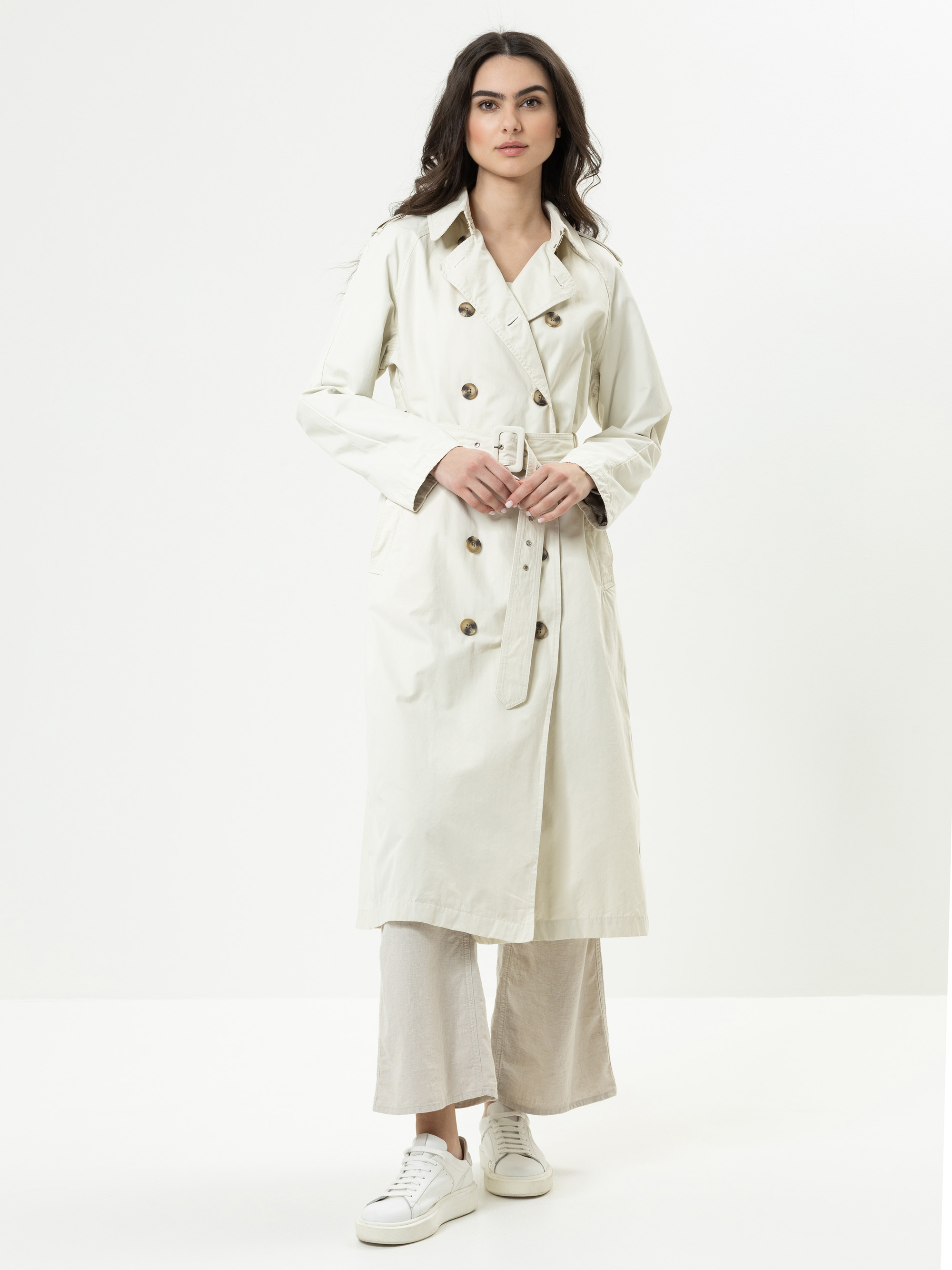 Eine Frau steht vor einem weißen Hintergrund und trägt einen langen, hellen Trenchcoat mit Gürtel, eine weit geschnittene Hose und weiße Turnschuhe. Sie hat langes dunkles Haar und einen neutralen Gesichtsausdruck.