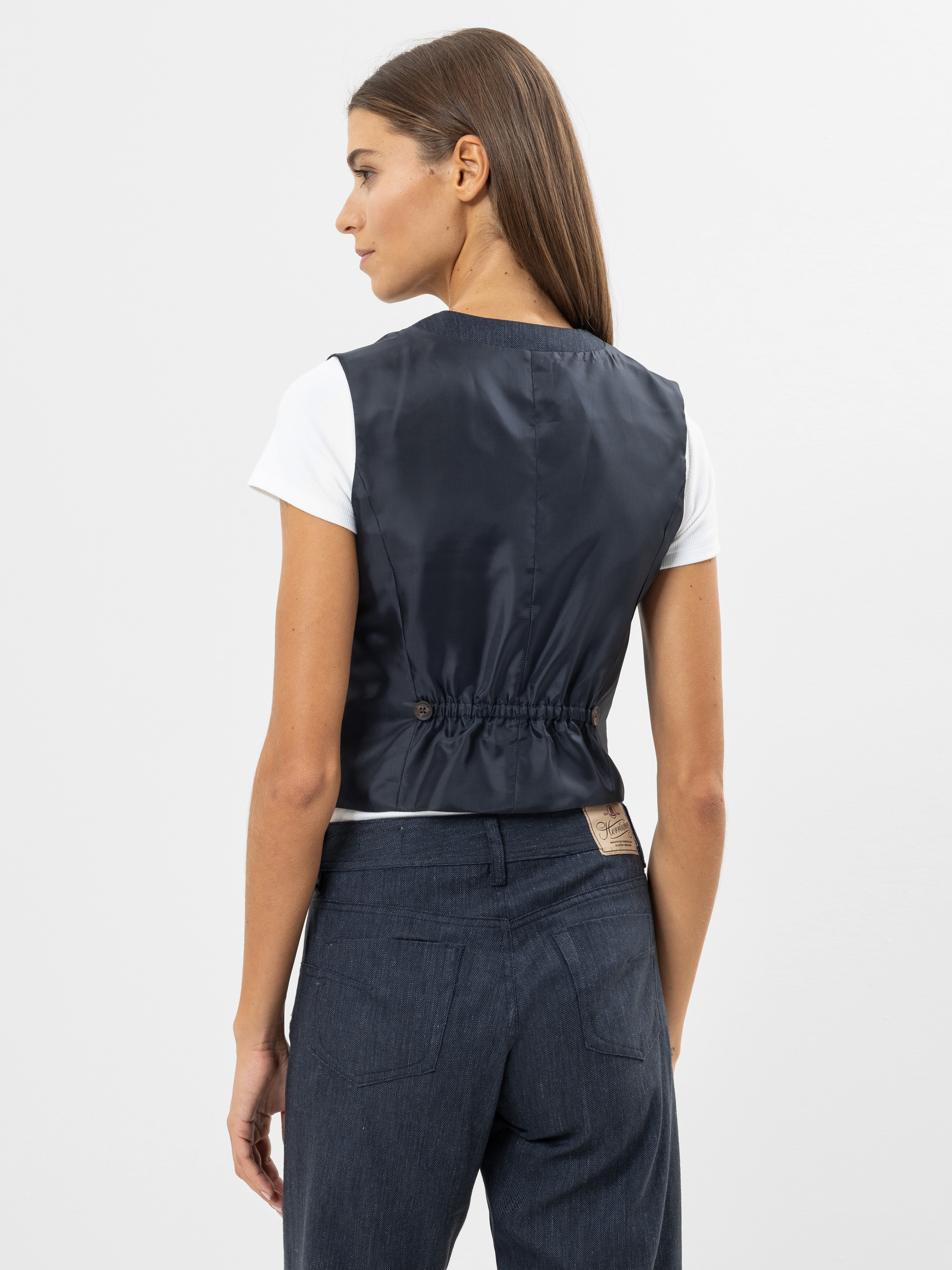 Eine Frau mit glattem braunem Haar wird von hinten gezeigt. Sie trägt eine marineblaue Satinweste über einem weißen kurzärmeligen Oberteil und dunkelblaue Jeans mit einem sichtbaren beigen Aufnäher am Bund.