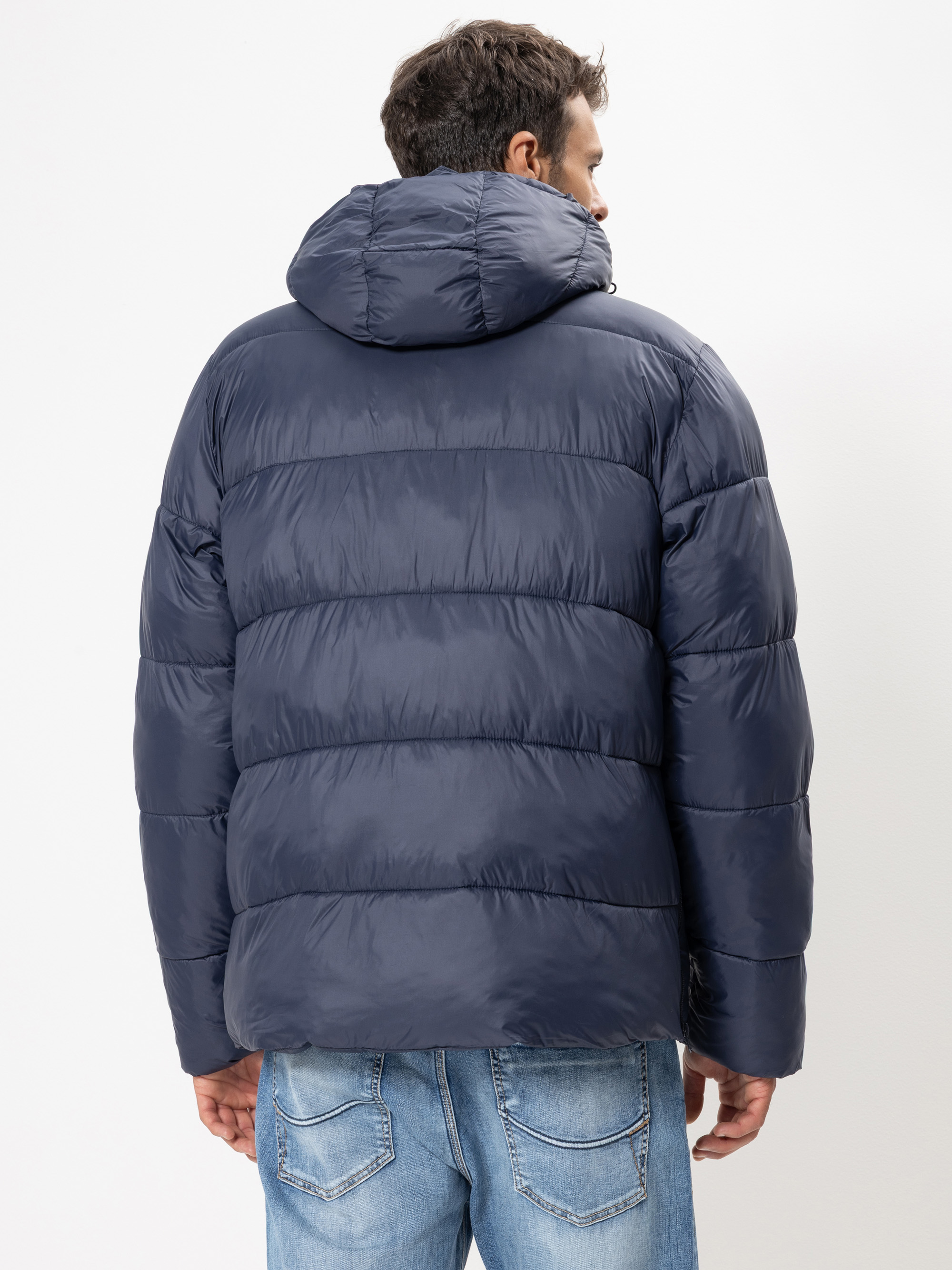 Ein Mann steht mit dem Rücken zur Kamera, trägt eine dunkelblaue Pufferjacke mit Kapuze und hellblaue Jeans vor einem schlichten weißen Hintergrund.