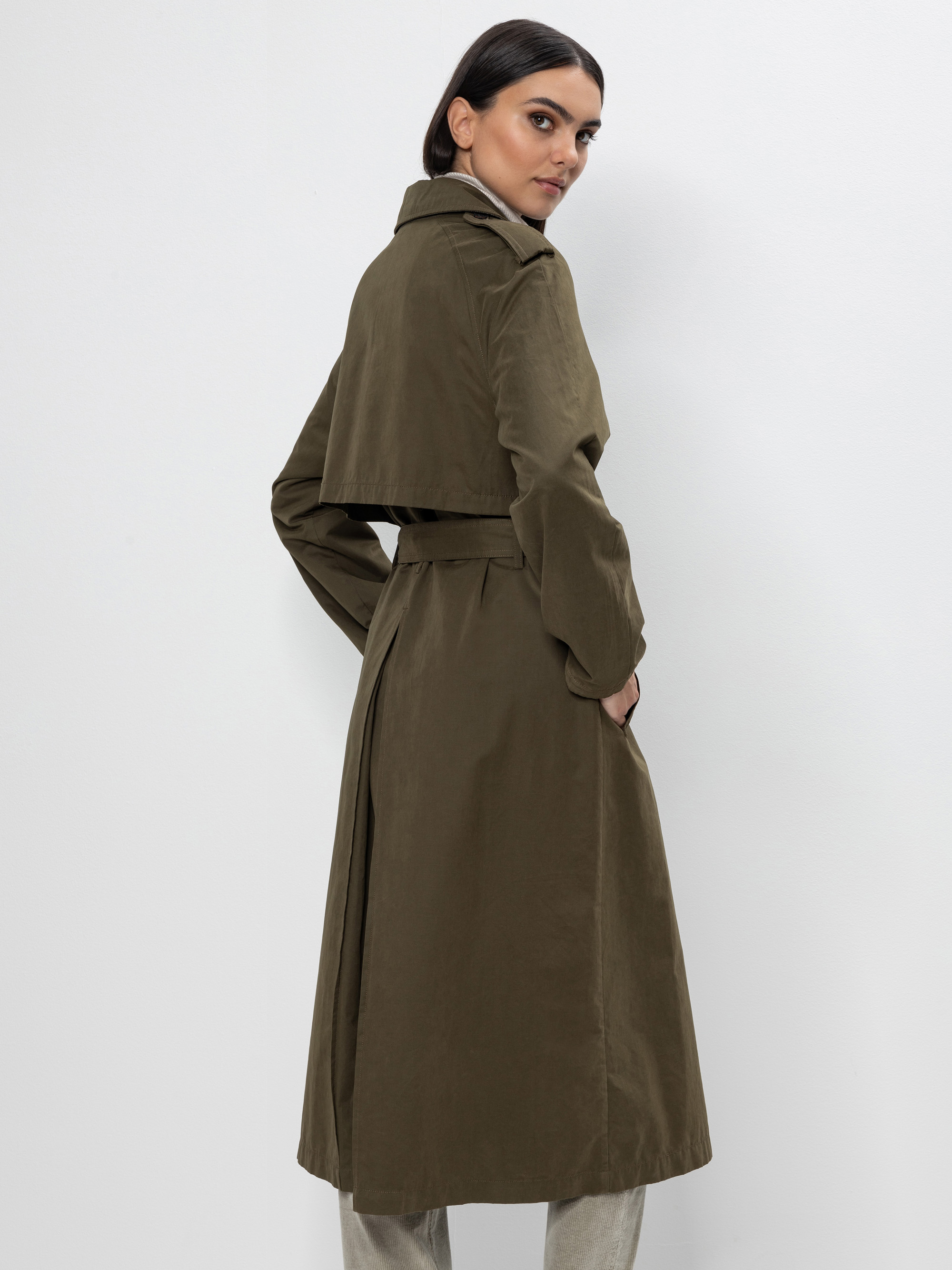 Eine Frau mit dunklem Haar, die einen olivgrünen Trenchcoat trägt, steht mit dem Rücken zugewandt, die Hände in den Manteltaschen, und blickt über ihre Schulter vor einem schlichten, hellen Hintergrund.
