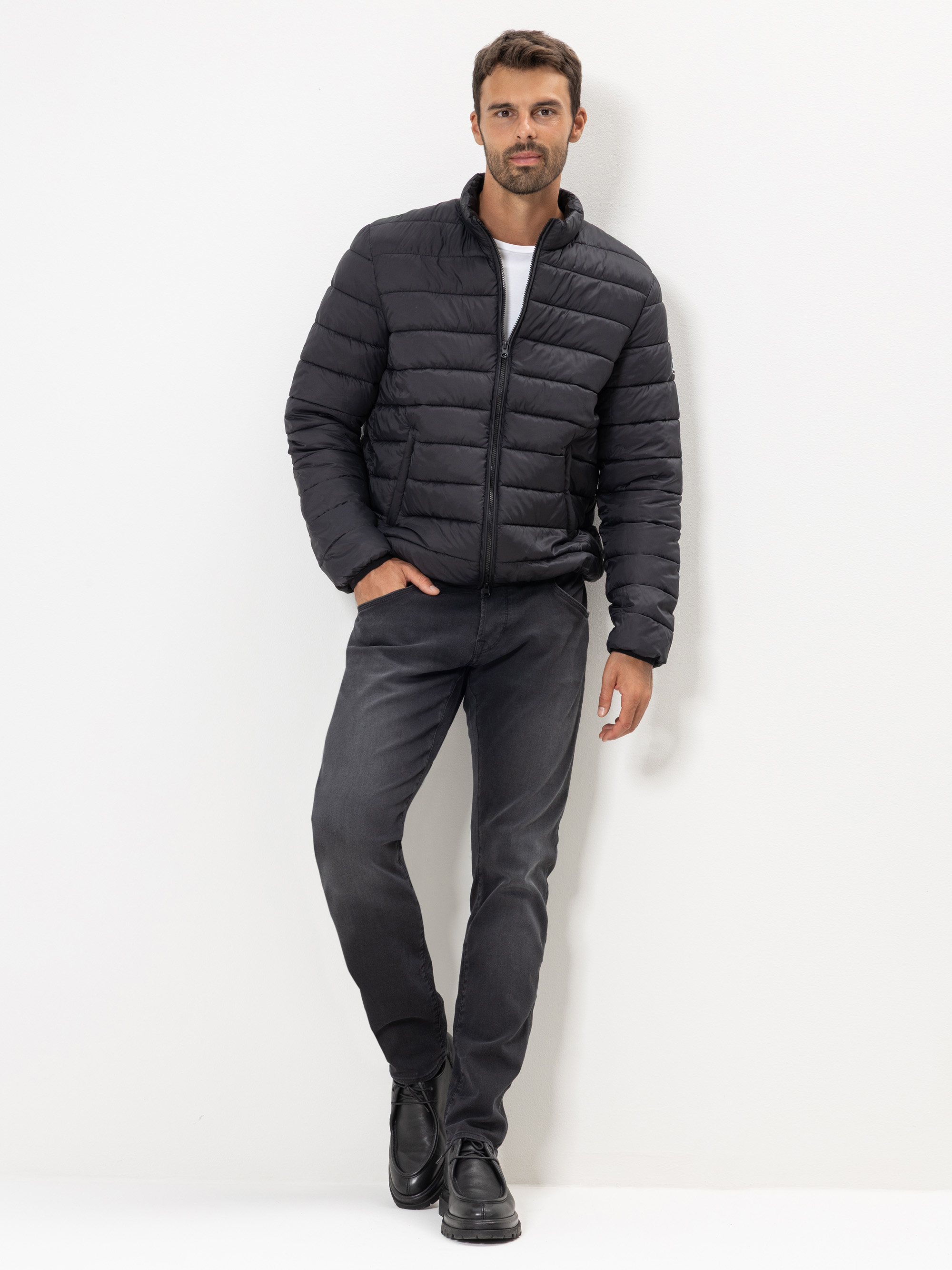 Ein Mann steht vor einem weißen Hintergrund und trägt eine schwarze Pufferjacke, ein weißes Hemd, dunkle Jeans und schwarze Schuhe. Er hat kurzes braunes Haar und einen gestutzten Bart, eine Hand in der Tasche und eine entspannte Körperhaltung.