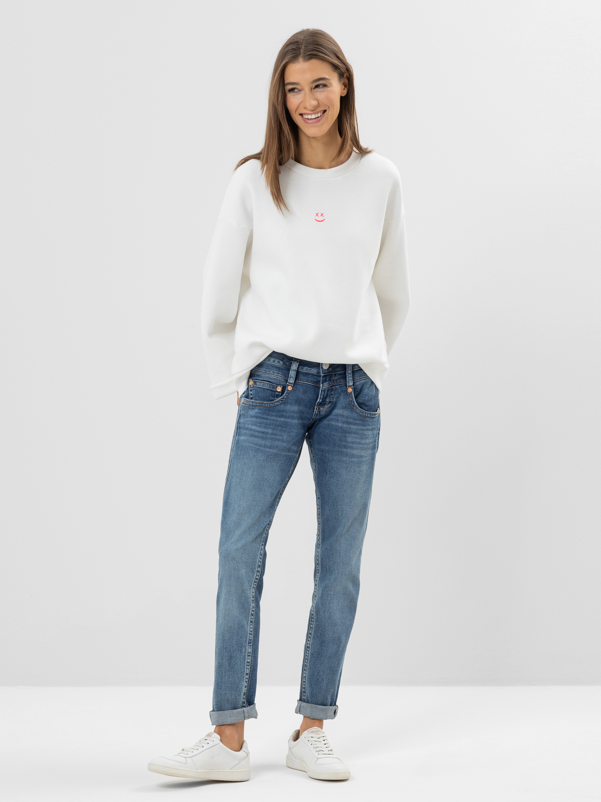 Eine Frau steht lächelnd vor einem schlichten Hintergrund und trägt ein weißes Sweatshirt mit einem kleinen rosa Muster, blaue Jeans und weiße Turnschuhe. Ihre Hände stecken in den Taschen und ihr Haar ist offen.