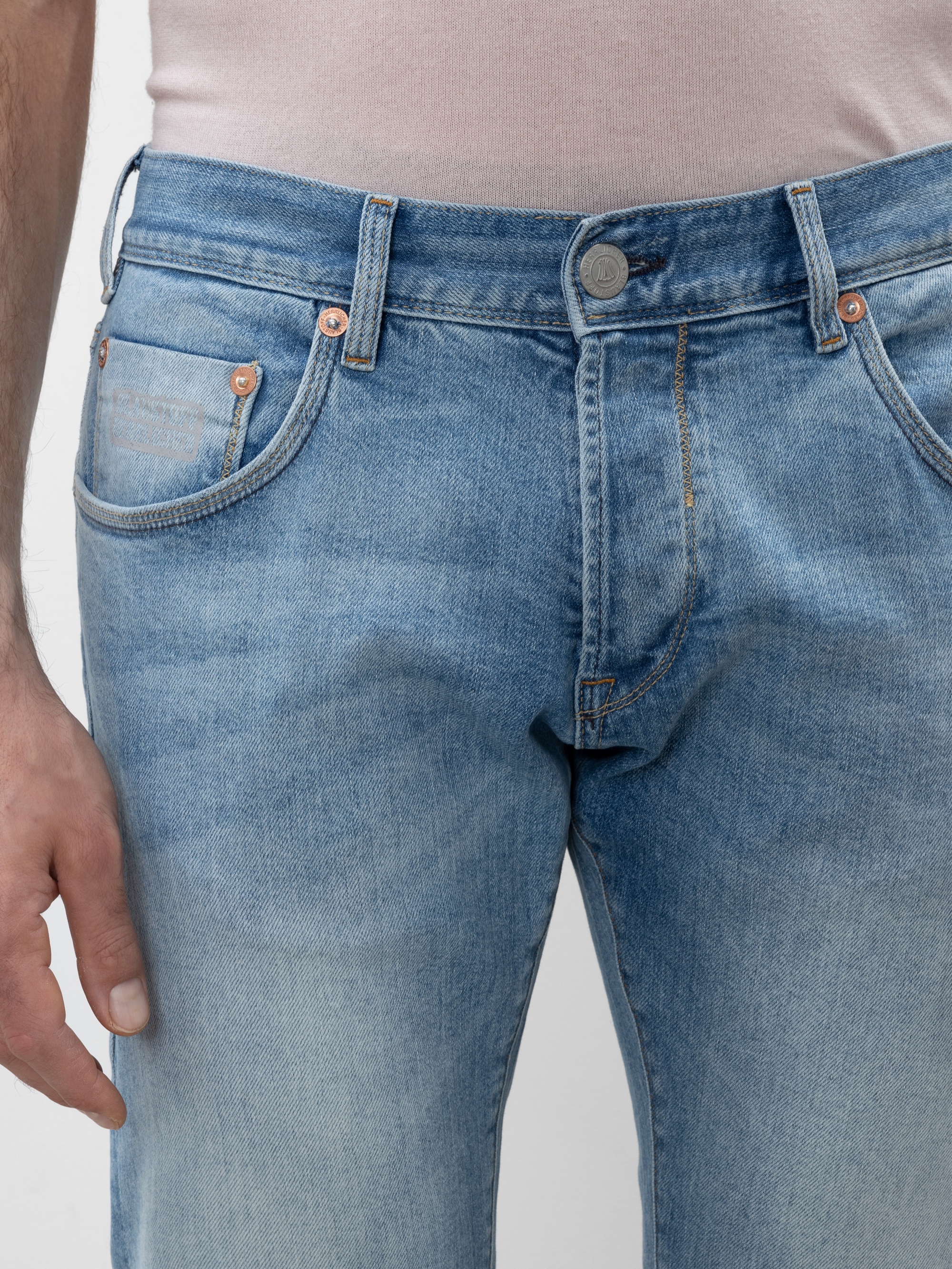 Eine Person, die eine hellblaue Jeans und ein hellrosa Hemd trägt, wobei der linke Arm entspannt an der Seite liegt. Das Bild konzentriert sich auf die Taille und den Oberschenkelbereich der Jeans.