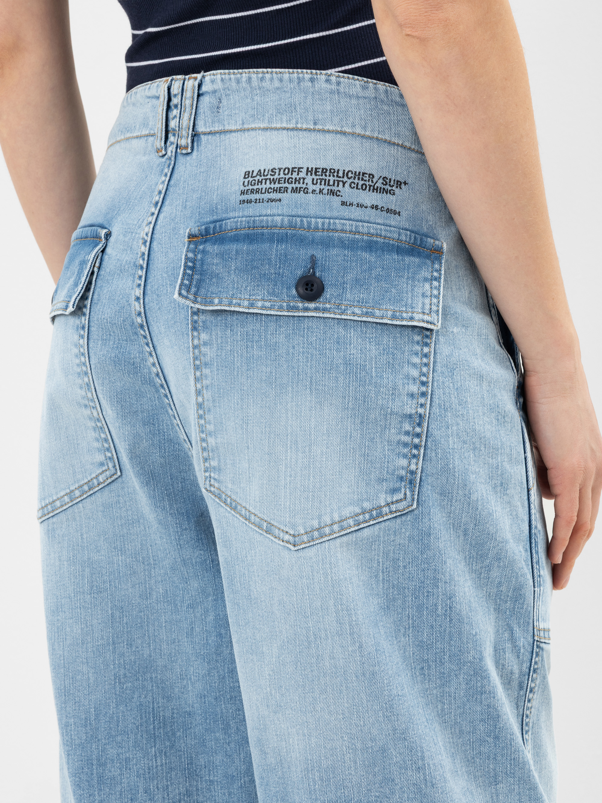 Nahaufnahme einer Person, die eine hellblaue Jeans mit weitem Bein und einer Gesäßtasche mit Knopfleiste und gedrucktem Text über der Tasche trägt. Die Person steht mit einer entspannten Hand an ihrer Seite.