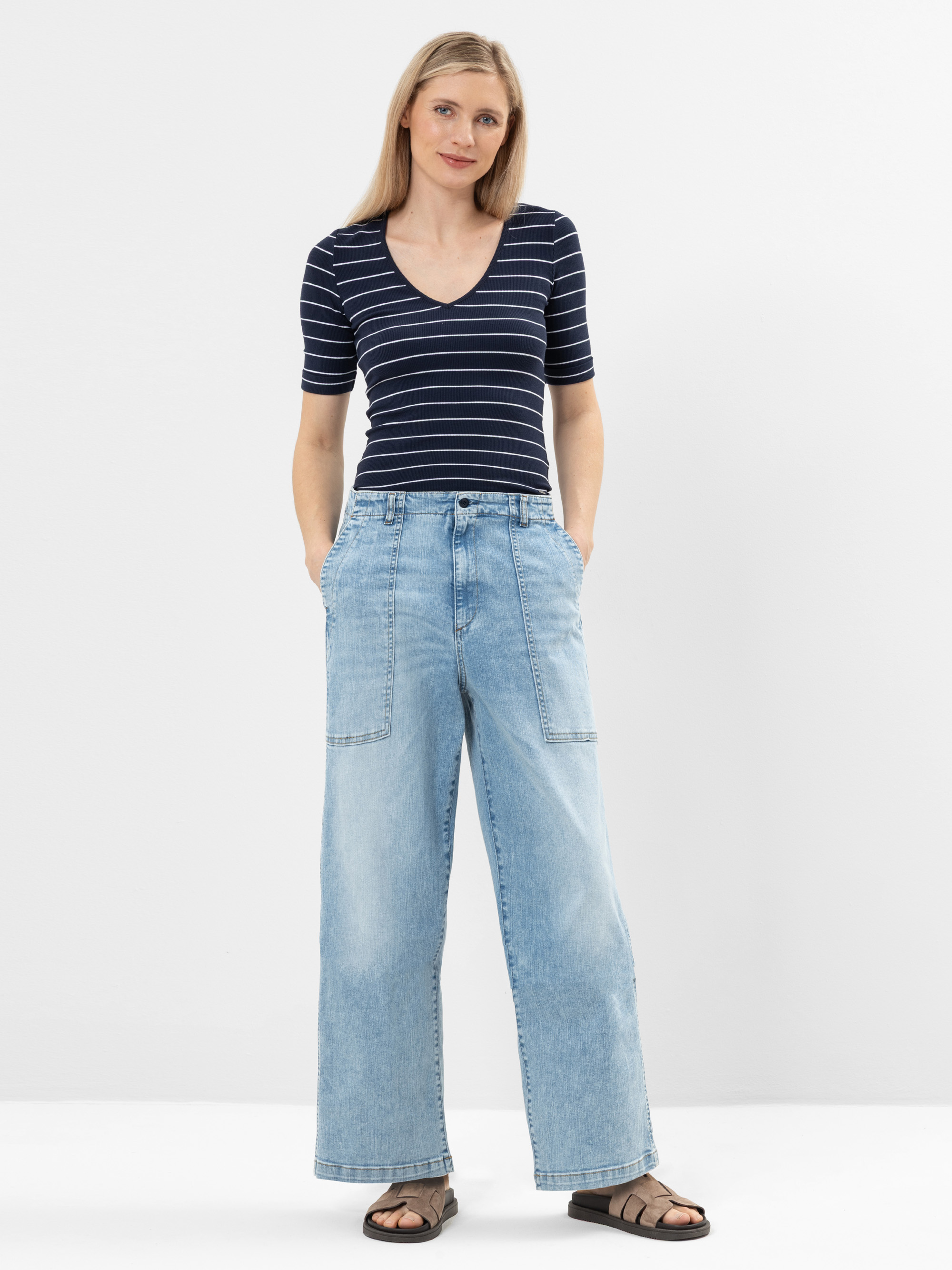 Eine Frau steht vor einem schlichten weißen Hintergrund und trägt ein marineblau gestreiftes, kurzärmeliges Oberteil, hellblaue, weit geschnittene Jeans mit großen Vordertaschen und dunkle Sandalen. Sie hat glattes blondes Haar und ihre Hände stecken