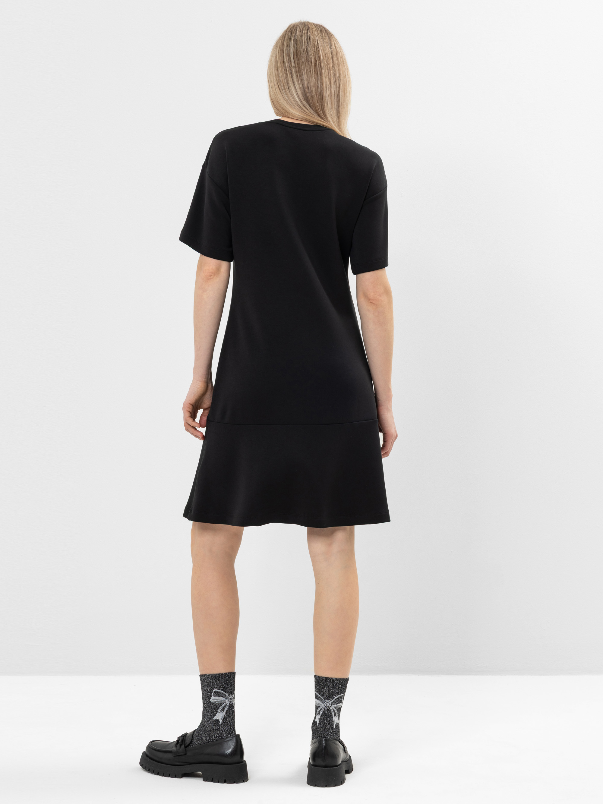 Eine Person mit glattem, blondem Haar trägt ein locker sitzendes schwarzes Kleid, gemusterte graue Socken und schwarze klobige Schuhe und steht mit dem Rücken zur Kamera vor einem schlichten weißen Hintergrund.