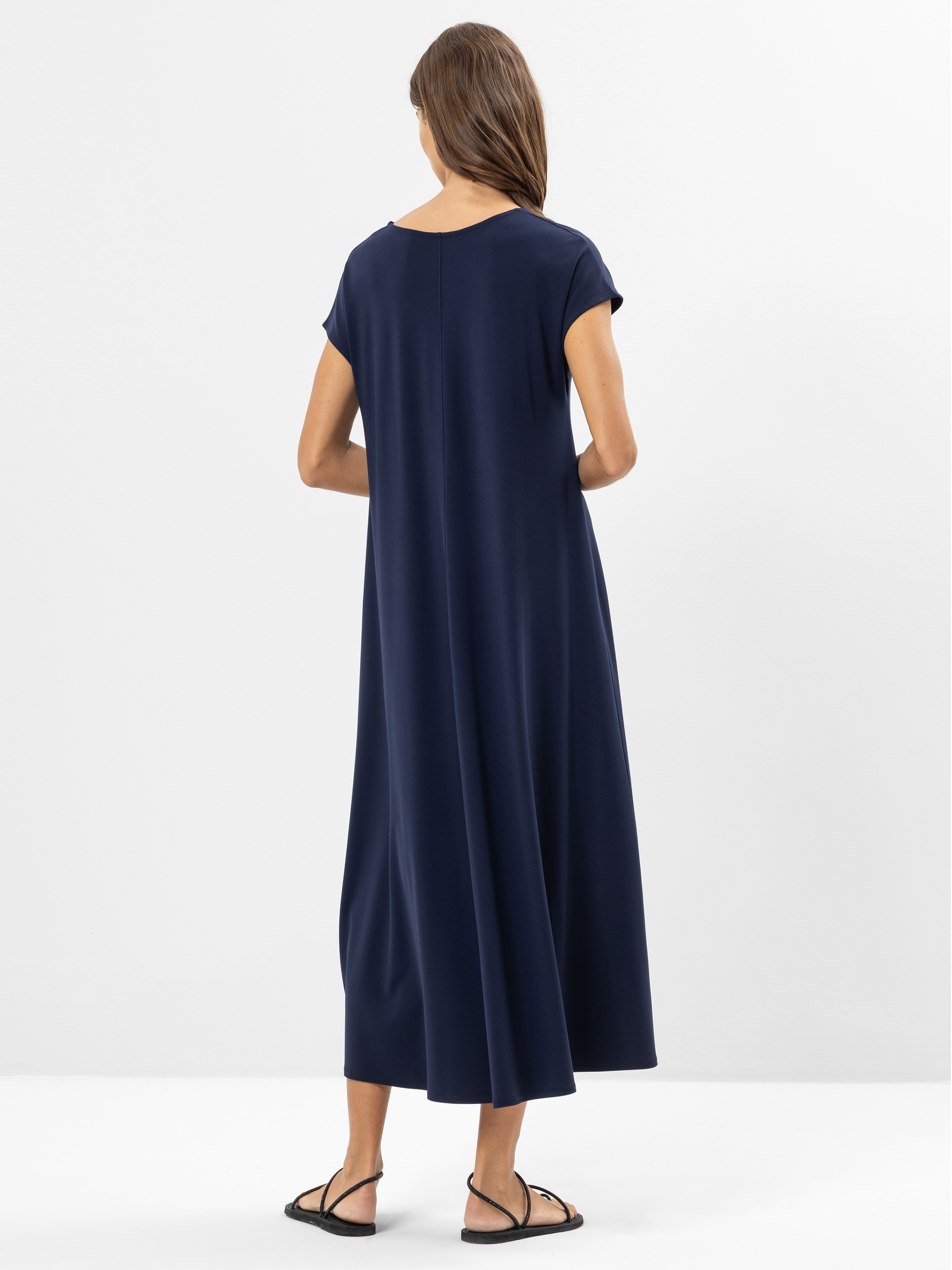 Eine Frau steht mit dem Gesicht zur Wand, trägt ein locker sitzendes, kurzärmeliges, knöchellanges, marineblaues Kleid und schwarze flache Sandalen vor einem schlichten weißen Hintergrund.