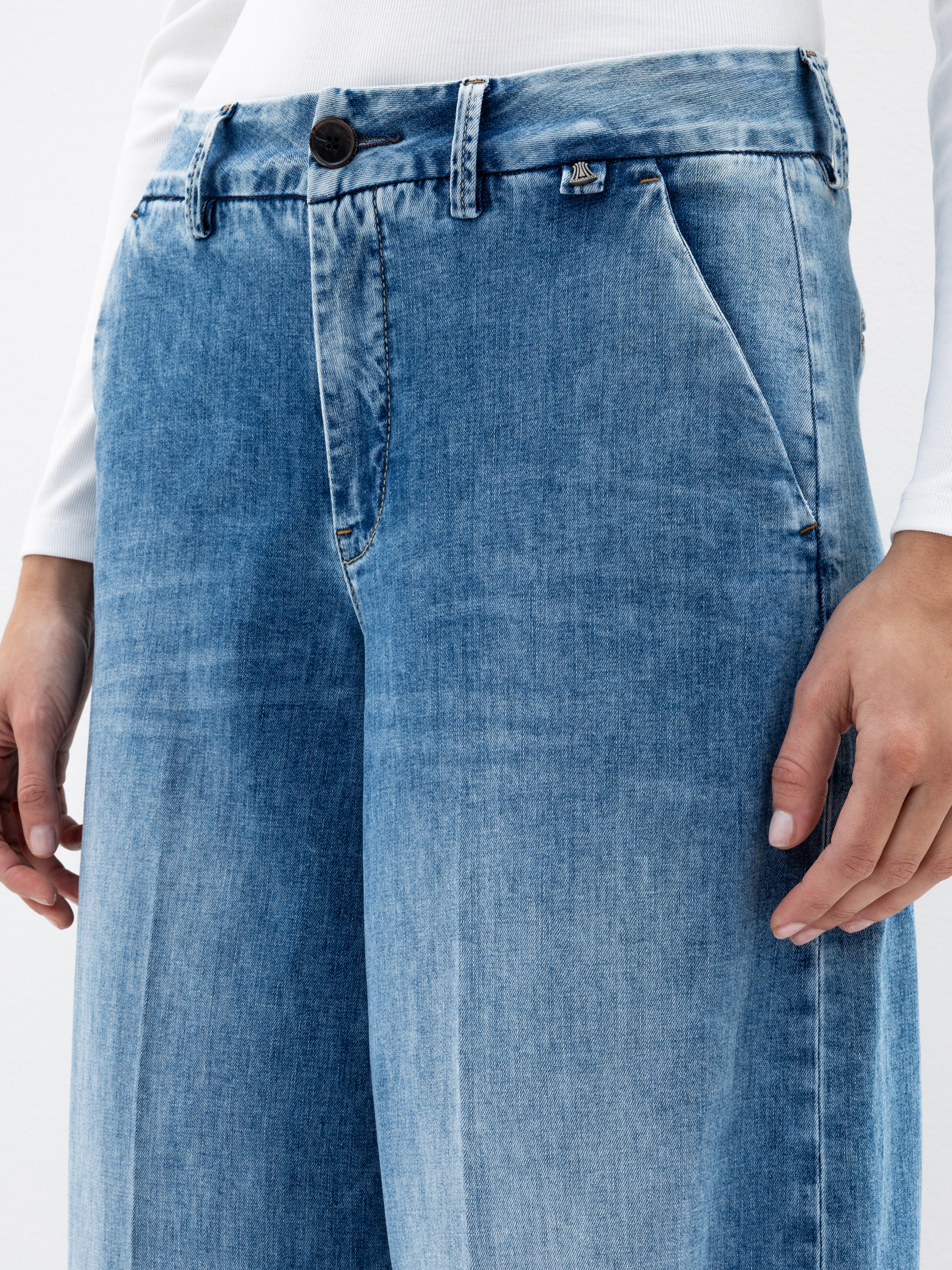 Eine Person, die hellblaue Jeans mit hoher Taille und weitem Bein und ein weißes langärmeliges Oberteil trägt, wobei die Hände an den Seiten der Hose ruhen. Der Schwerpunkt des Fotos liegt auf der Taille und den Oberschenkeln.
