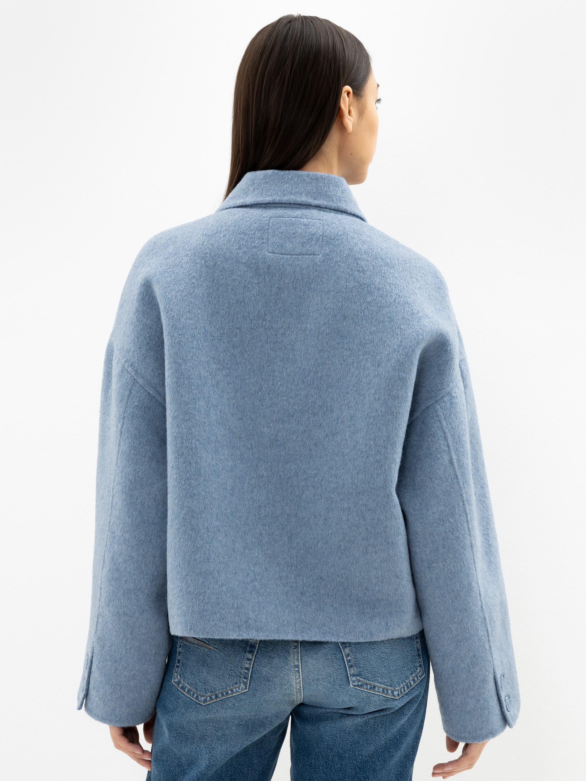 Eine Person mit langen dunklen Haaren, von hinten gesehen, trägt einen hellblauen Wollmantel und blaue Jeans und steht vor einem schlichten weißen Hintergrund.