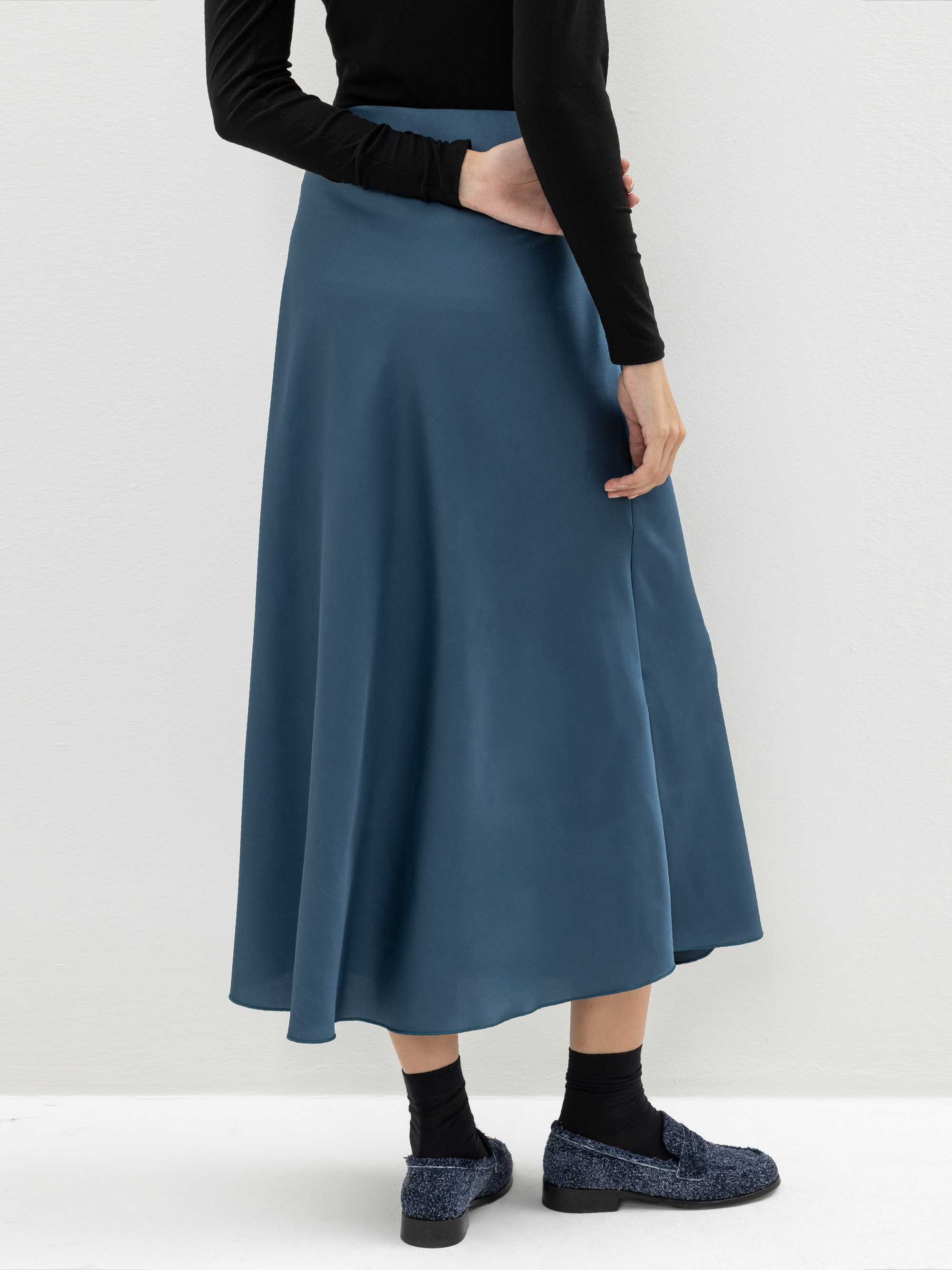 Eine Person, die einen langen, fließenden blauen Rock, ein schwarzes, langärmeliges Oberteil, schwarze Socken und blaue Halbschuhe trägt, steht vor einer schlichten, hellen Wand und hat eine Hand auf die Hüfte gestützt.