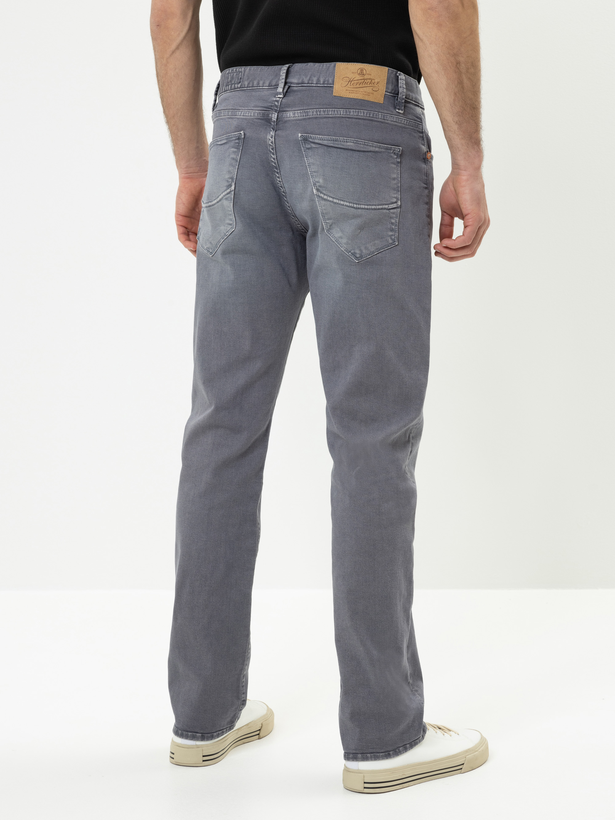 Ein Mann steht mit dem Gesicht zur Wand und trägt eine hellgraue Jeans, ein schwarzes Hemd und weiße Turnschuhe. Die Jeans hat sichtbare Gesäßtaschen und einen hellbraunen Lederaufnäher am Bund. Der Hintergrund ist einfarbig weiß.