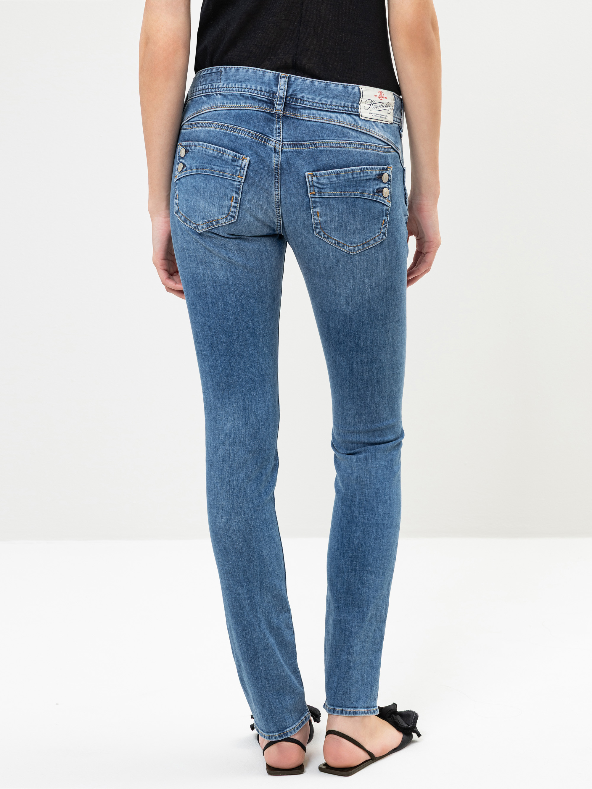 Eine Person, die blaue Skinny Jeans und schwarze Sandalen trägt, wird von hinten gezeigt und steht vor einem schlichten hellen Hintergrund. Die Jeans hat zwei Gesäßtaschen mit Knopfklappen.