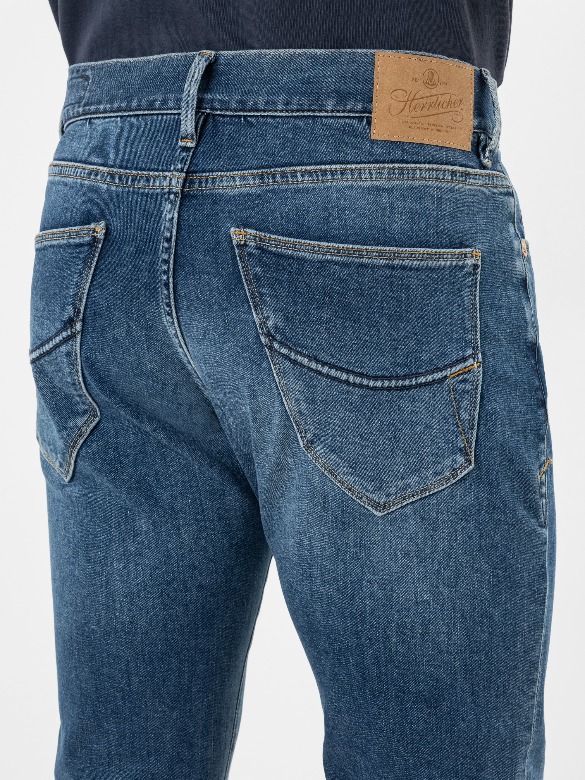 Nahaufnahme des Rückens einer Person, die eine blaue Denim-Jeans mit zwei Gesäßtaschen und einem braunen Lederetikett am Bund trägt. Die Jeans hat sichtbare Nähte und leichte Ausbleichungen auf dem Stoff.