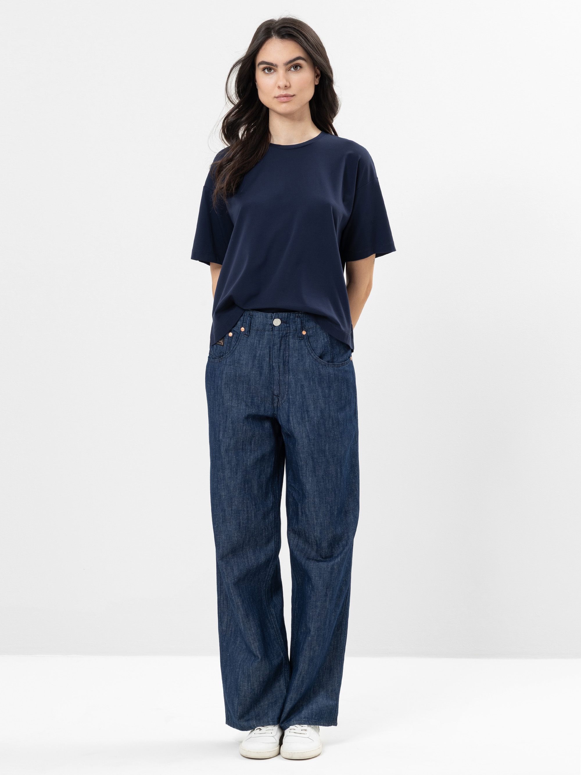 Eine Frau steht vor einem einfarbig weißen Hintergrund und trägt ein marineblaues T-Shirt, eine locker sitzende Bluejeans und weiße Turnschuhe. Sie hat die Hände hinter dem Rücken verschränkt und trägt langes, dunkles Haar.