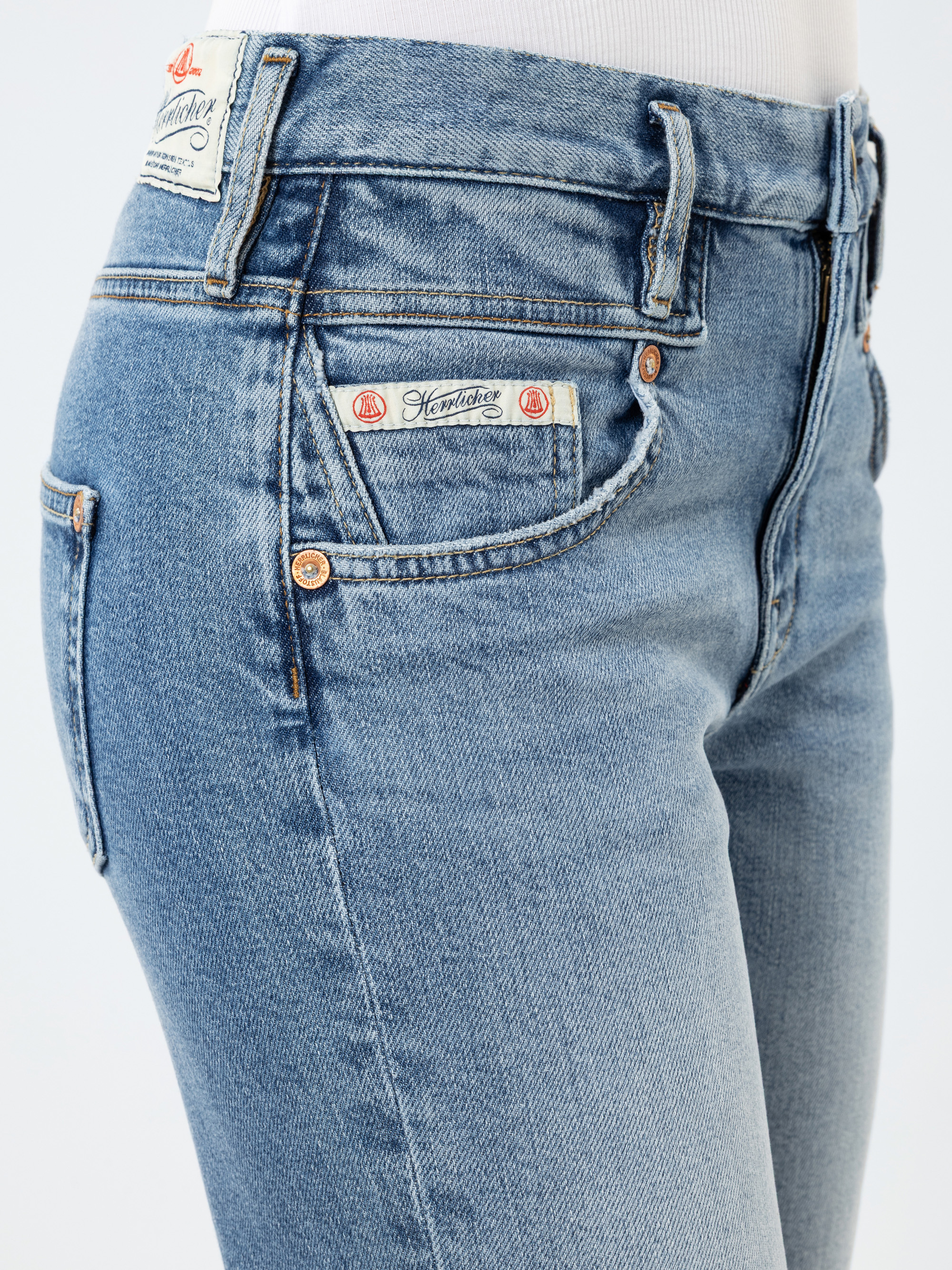 Nahaufnahme einer Person, die eine hellblaue Jeans mit hoher Taille und sichtbaren Taschen, Gürtelschlaufen und einem aufgenähten Markenetikett über der rechten Vordertasche trägt. Die Person trägt außerdem ein weißes Oberteil, das in die Jeans ges