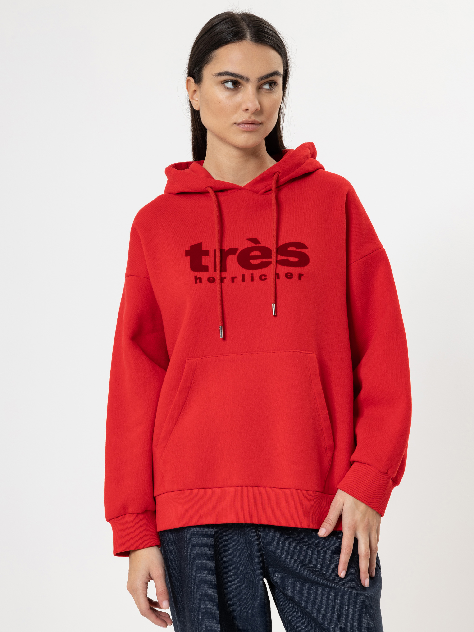 Eine Frau mit langen dunklen Haaren trägt einen leuchtend roten Kapuzenpulli, auf dem in schwarzer Schrift très und terrilicer steht. Sie steht vor einem schlichten weißen Hintergrund, hat die linke Hand in der Tasche und schaut zur Seite.