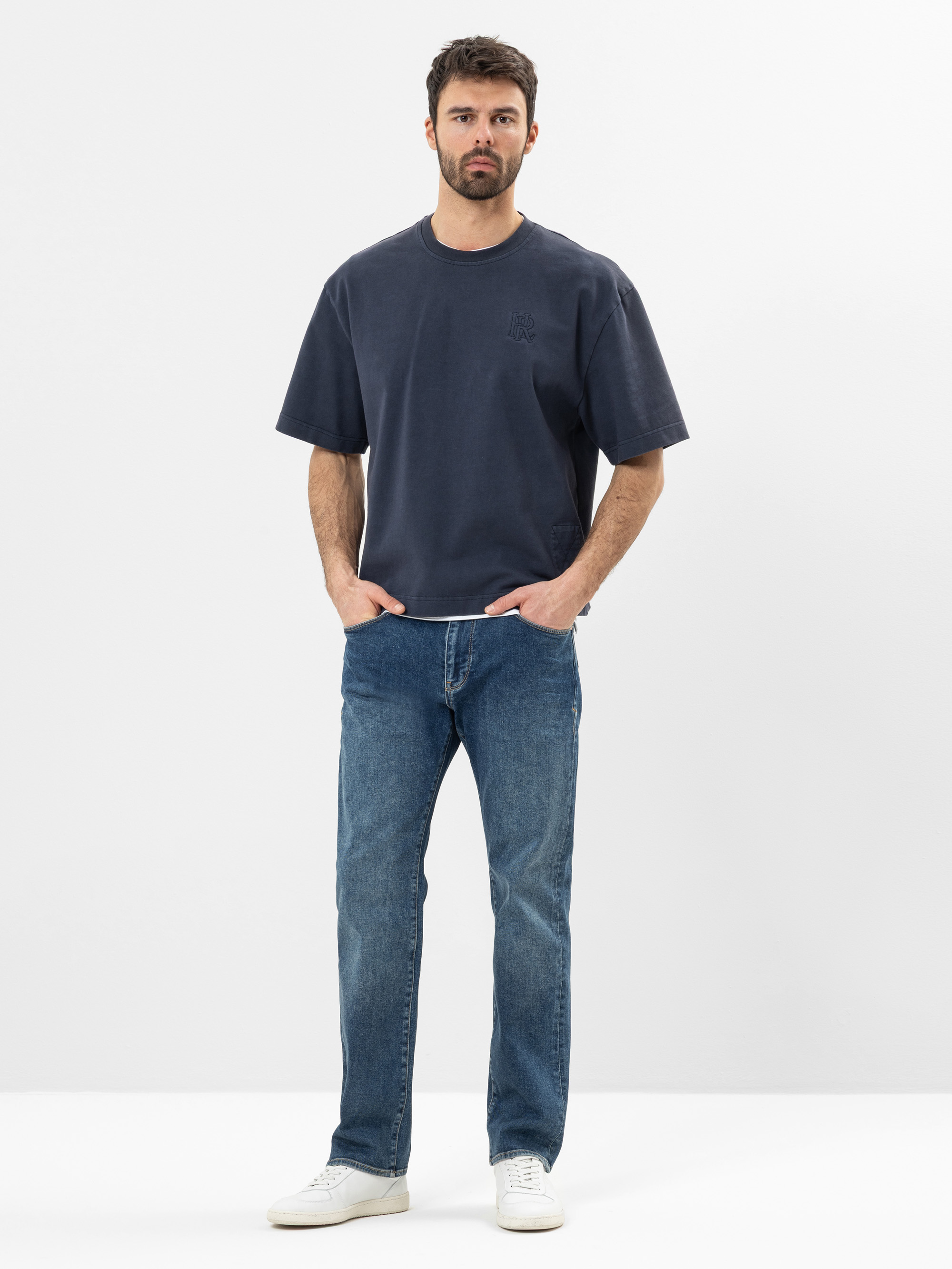 Ein Mann steht vor einem schlichten weißen Hintergrund, trägt ein locker sitzendes dunkelblaues T-Shirt, blaue Jeans und weiße Turnschuhe, die Hände in den Taschen und einen neutralen Gesichtsausdruck.