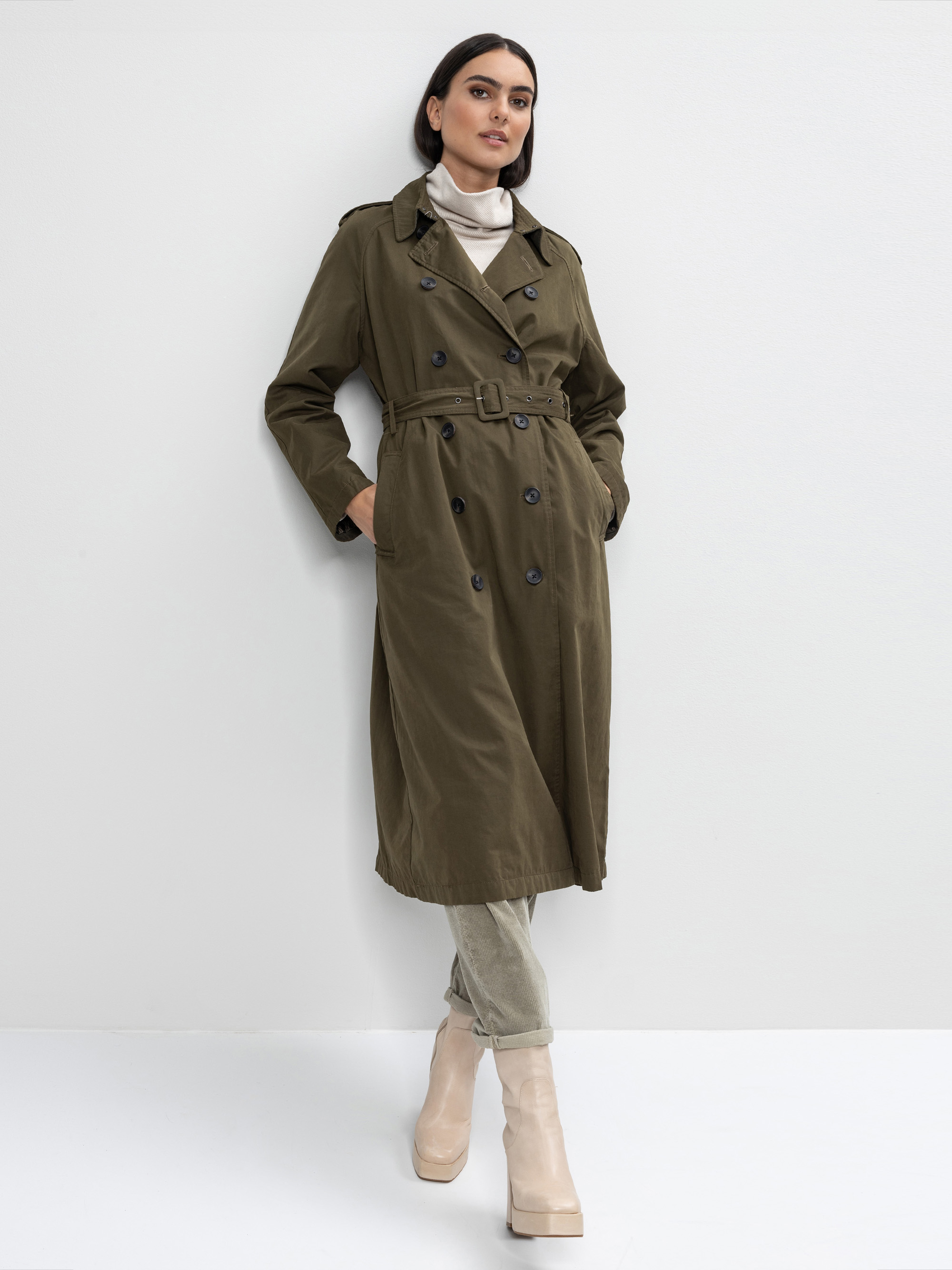Eine Frau, die einen langen olivgrünen Trenchcoat, einen hellbeigen Rollkragenpullover, eine helle Hose und beigefarbene Plateaustiefel trägt, steht vor einer schlichten weißen Wand, hat die Hände in den Taschen und schaut leicht zur Seite.