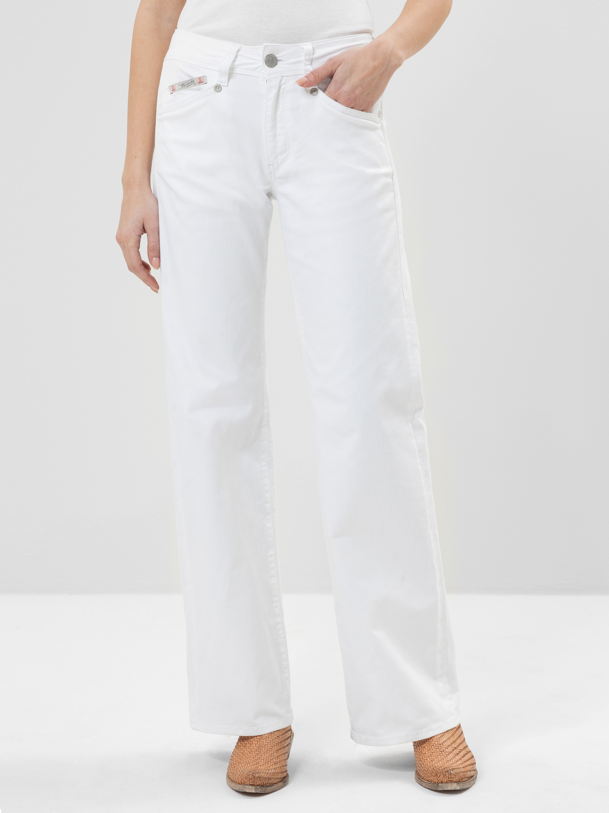 Eine Person, die eine weiße Jeans mit hoher Taille und weitem Bein, ein weißes Oberteil und hellbraune Webschuhe trägt, steht vor einem schlichten hellen Hintergrund.
