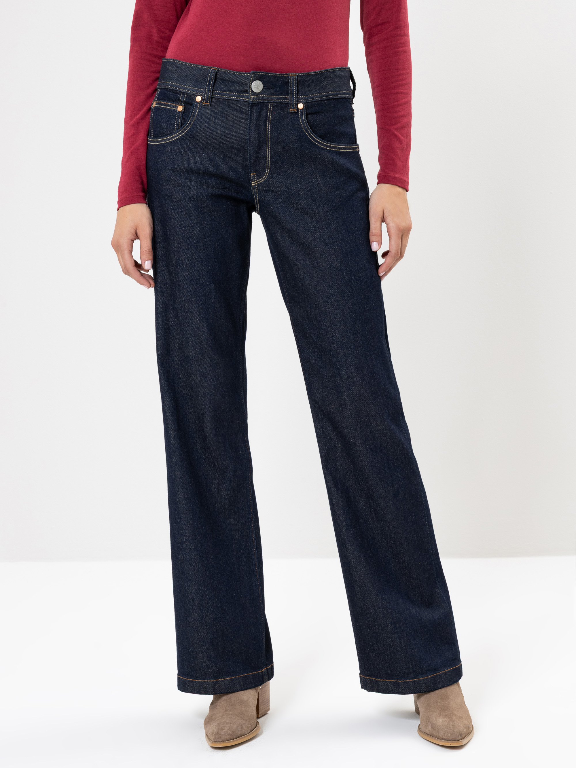 Person, die dunkelblaue Jeans mit weitem Bein, ein langärmeliges rotes Hemd und beigefarbene Ankle Boots trägt und vor einem schlichten weißen Hintergrund steht. Nur der Unterkörper ist sichtbar.