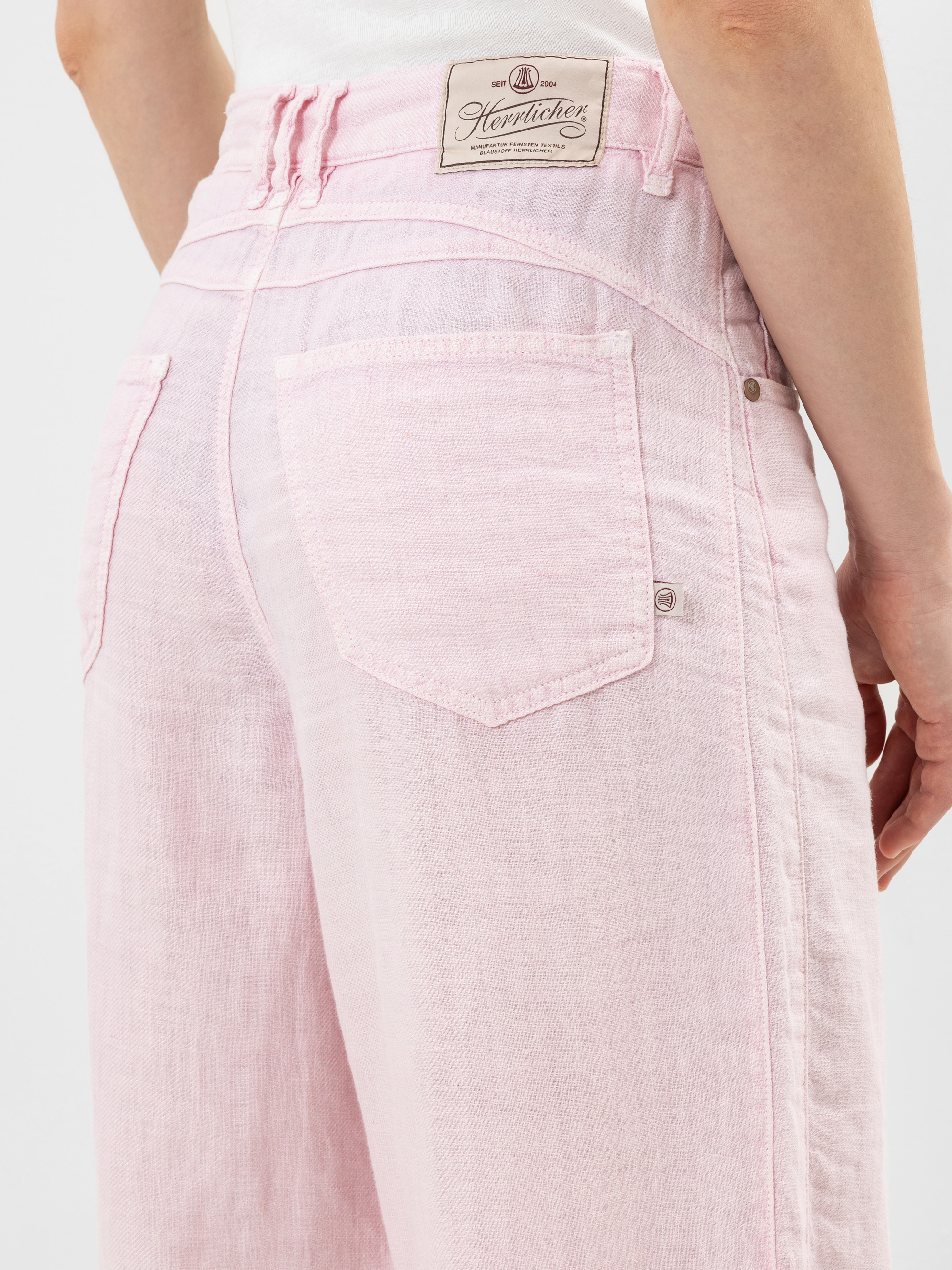 Nahaufnahme einer Person, die eine hellrosa High-Waisted-Jeans mit einer sichtbaren Gesäßtasche und einem weißen Herrlicher-Markenetikett am Bund trägt. Die Person trägt ein weißes Oberteil, das in die Jeans gesteckt wird.