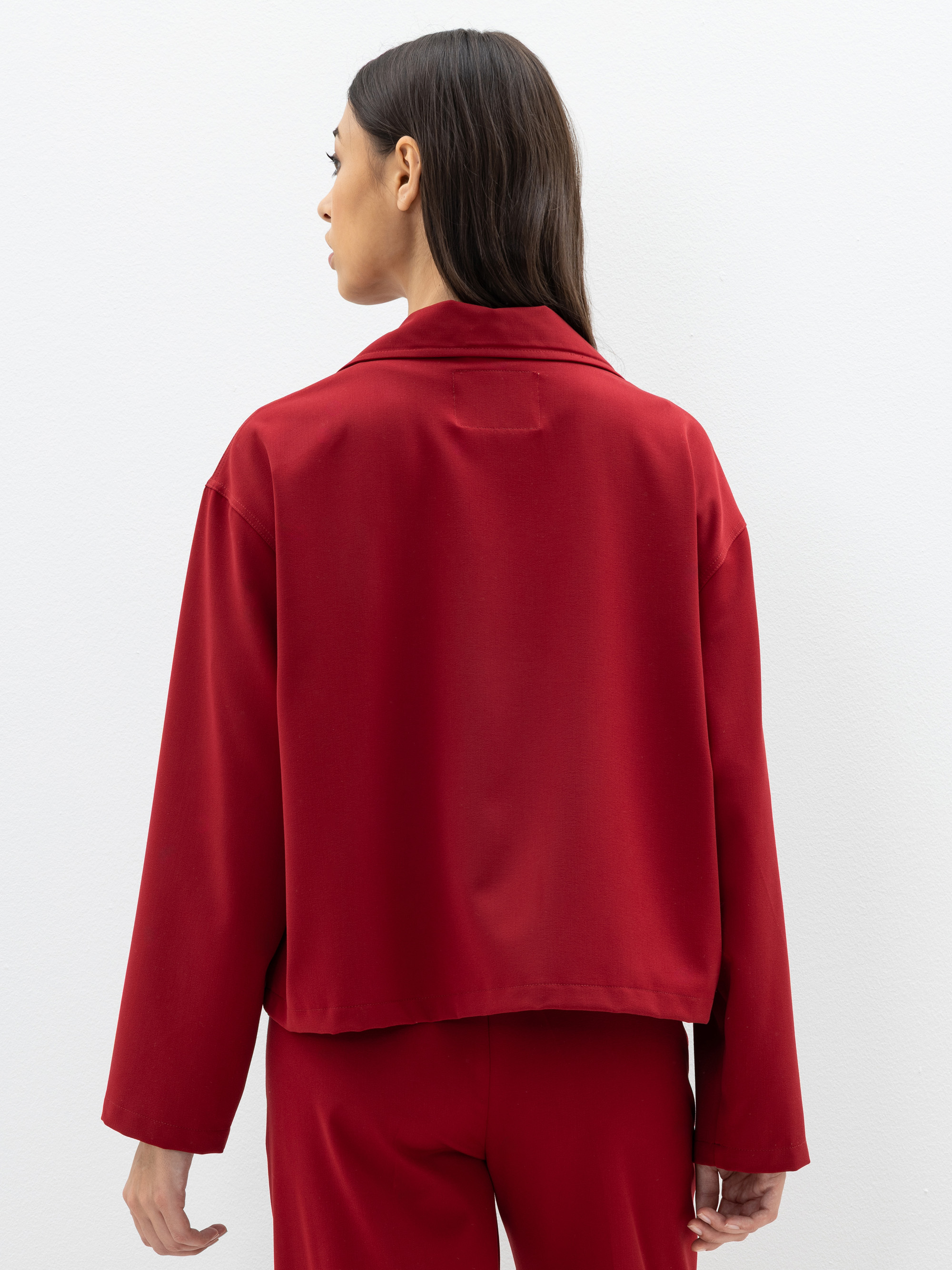 Eine Frau mit langen dunklen Haaren steht mit dem Gesicht zur Wand und trägt einen locker sitzenden roten Blazer und eine dazu passende Hose vor einem schlichten weißen Hintergrund.