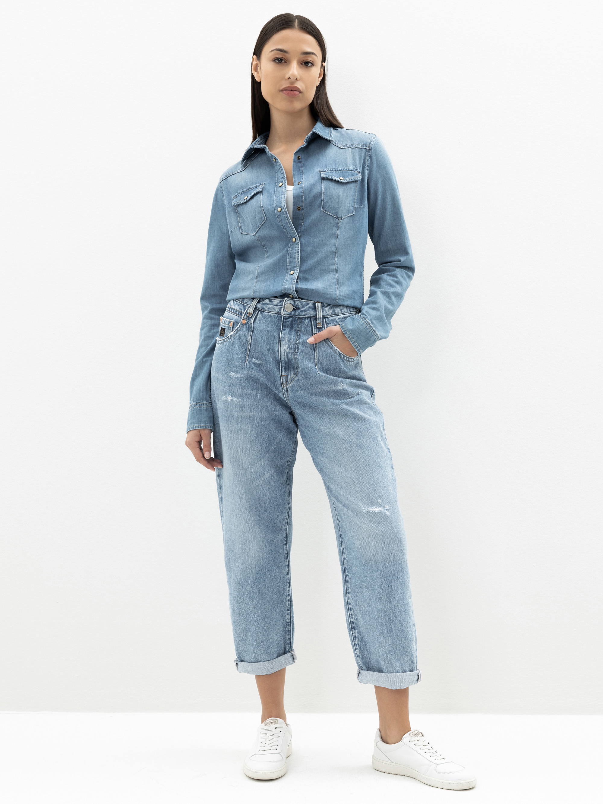 Eine Frau steht vor einem schlichten weißen Hintergrund und trägt ein hellblaues Jeanshemd mit Knopfleiste, das in eine Jeans mit hoher Taille und aufgerollten Manschetten gesteckt wird, sowie weiße Turnschuhe. Sie hat glattes, dunkles Haar und einen n