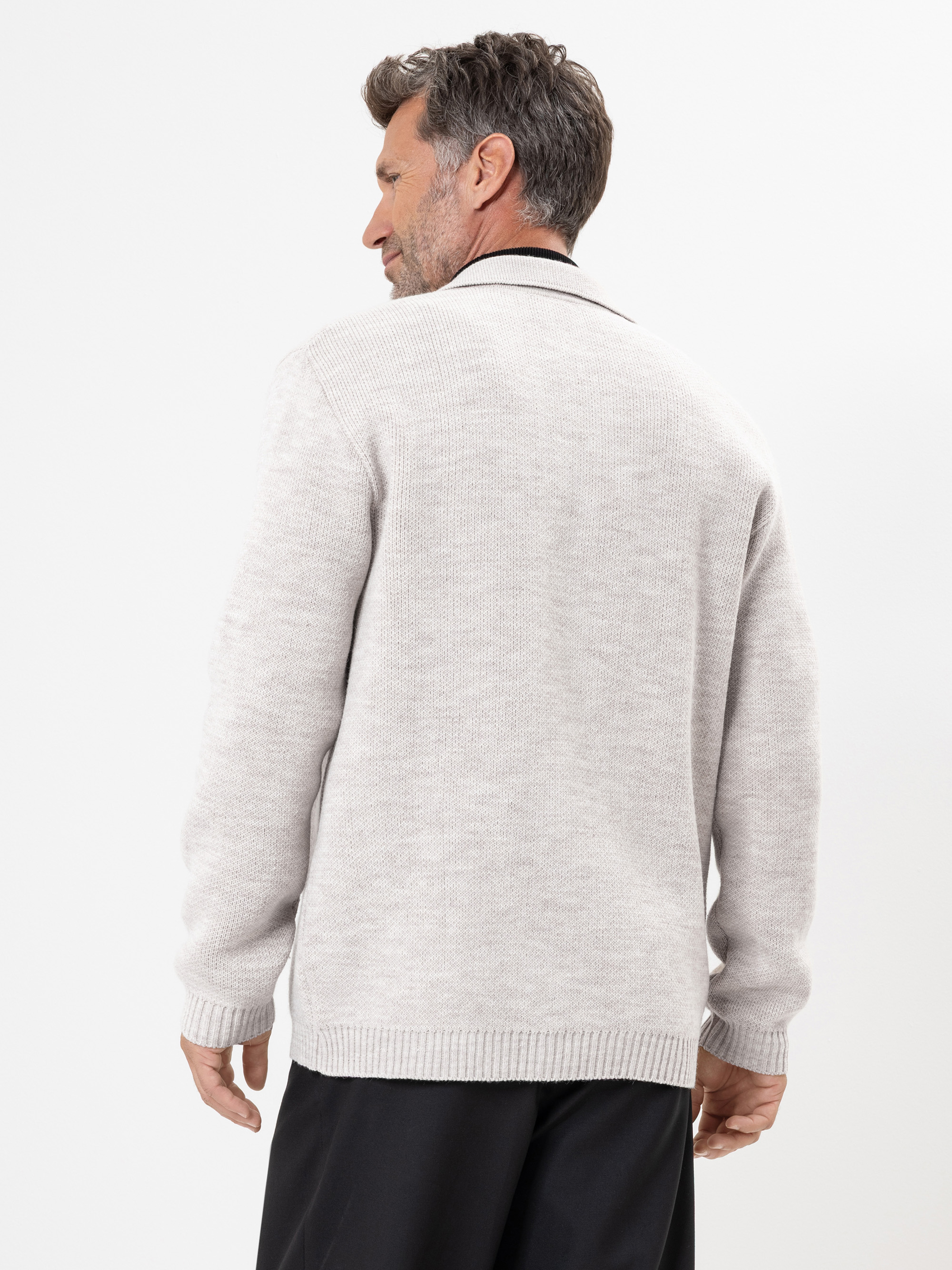 Ein Mann mit kurzen, dunklen Haaren und etwas grauem Haar steht mit dem Gesicht zur Wand. Er trägt eine hellbeige Strickjacke mit langen Ärmeln und gerippten Rändern, dazu eine schwarze Hose vor einem schlichten weißen Hintergrund.