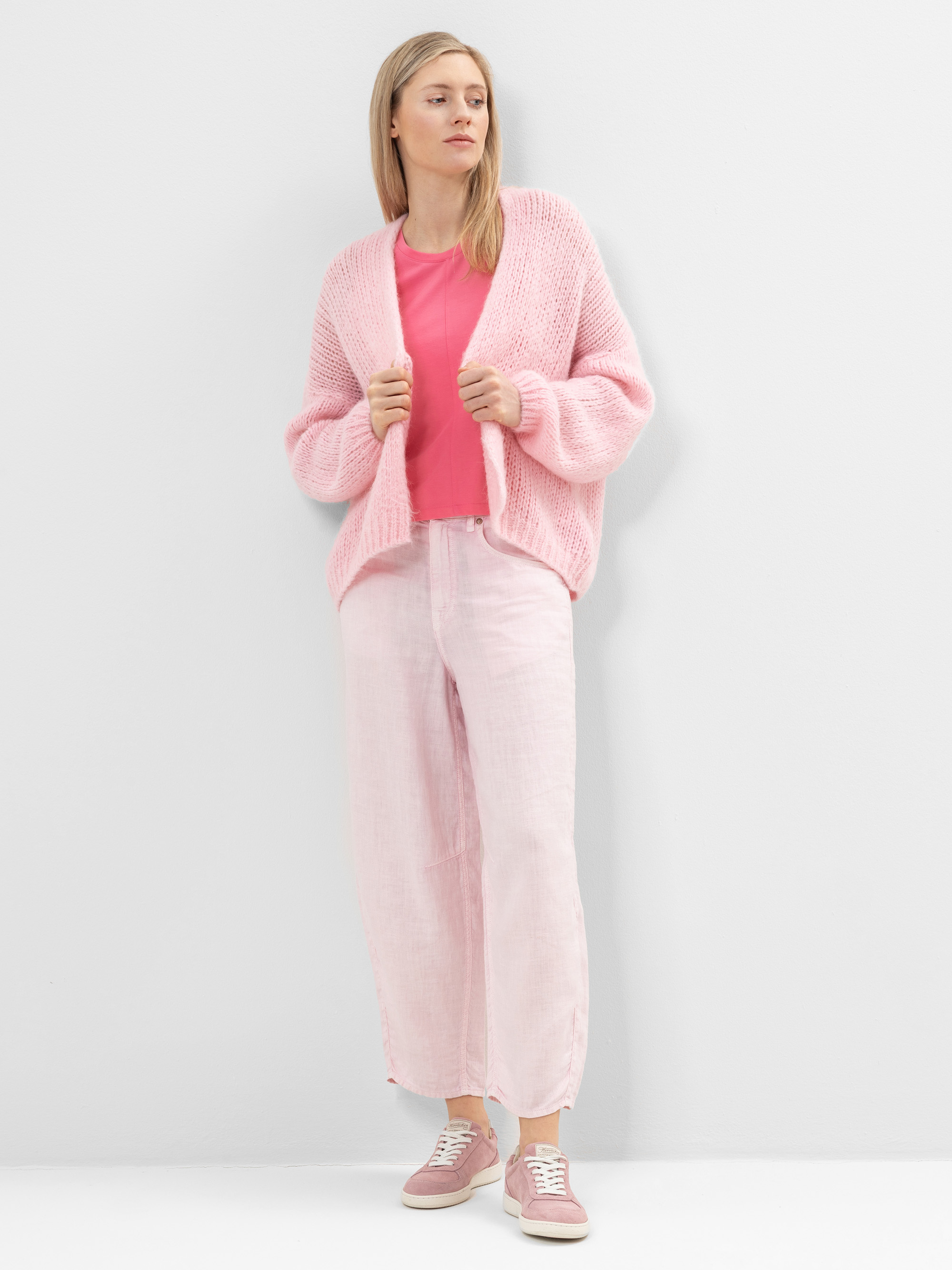 Eine Frau steht vor einer weißen Wand und trägt eine rosafarbene Strickjacke, ein rosafarbenes T-Shirt, eine hellrosa Hose mit weitem Bein und rosa Turnschuhe. Sie hat glattes blondes Haar und einen neutralen Gesichtsausdruck.