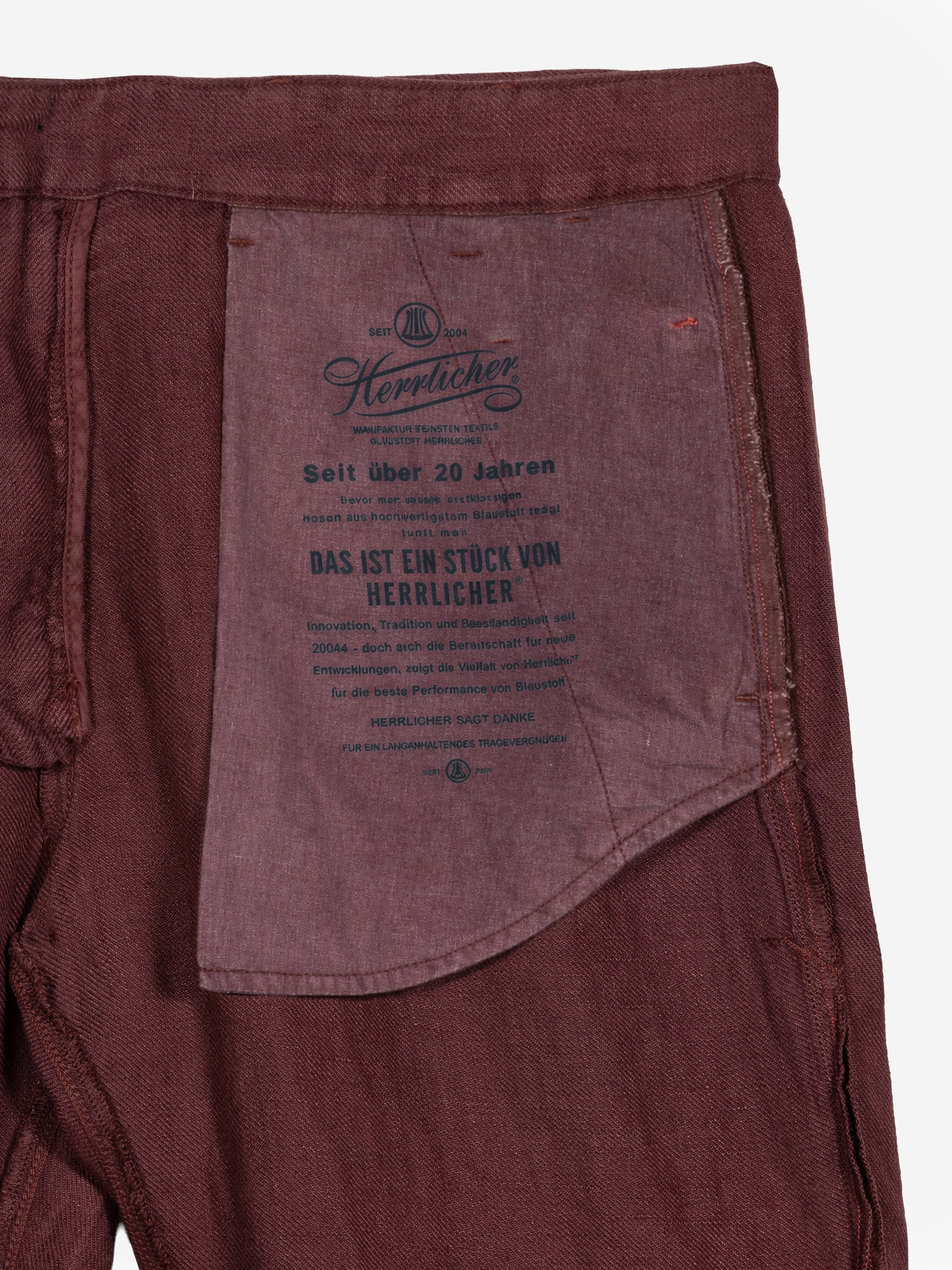 Nahaufnahme der Innenseite einer kastanienbraunen Hose, die eine große eingenähte Tasche mit aufgedrucktem deutschen Text und der Marke Herrlicher auf dem Innenfutter der Tasche zeigt. Die Hose ist flach vor einem weißen Hintergrund ausgebreitet.