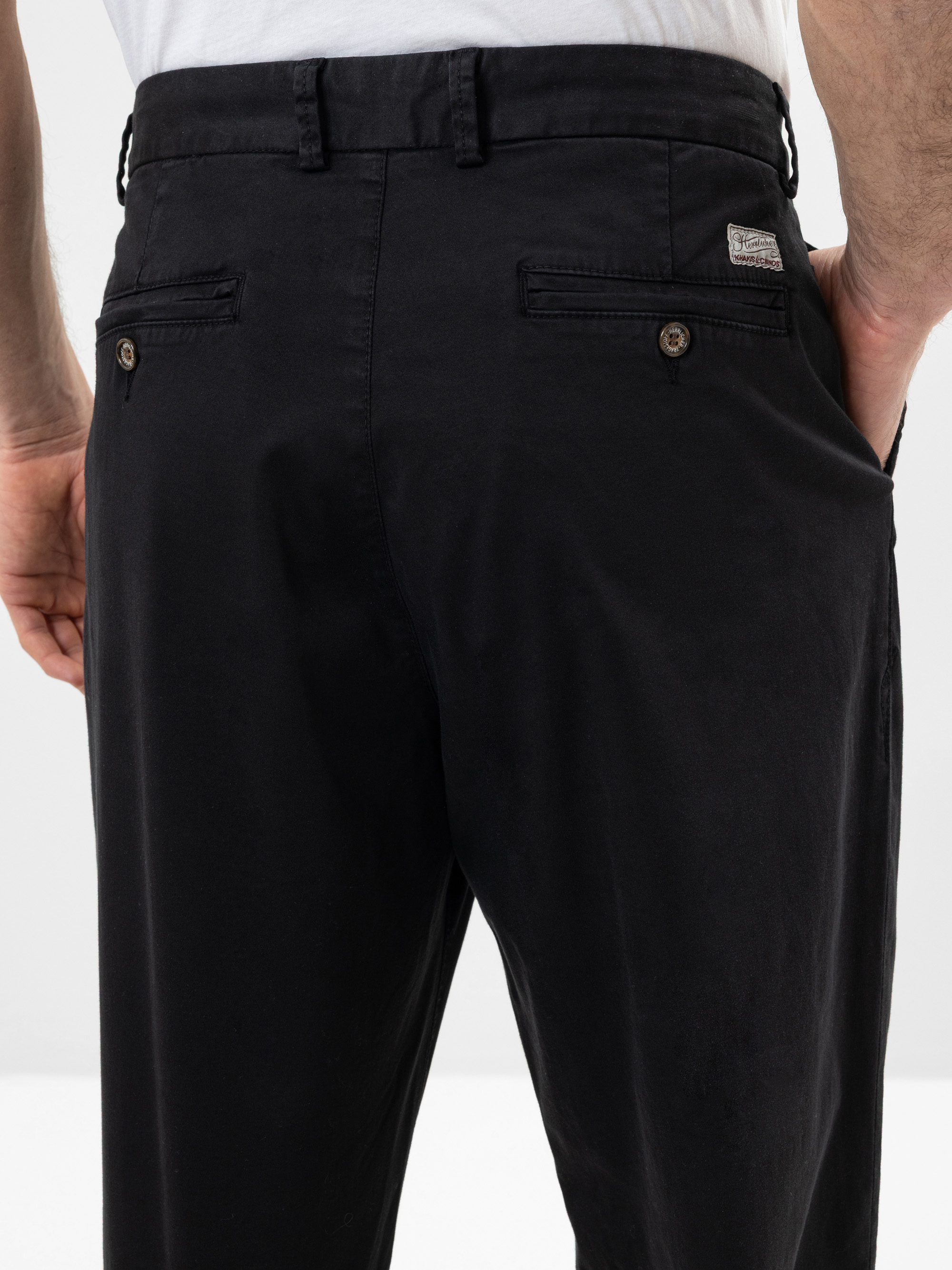 Nahaufnahme einer Person, die eine schwarze Anzughose und ein weißes Hemd trägt, wobei die Gesäßtaschen mit Knopfverschluss und Gürtelschlaufen zu sehen sind. Die Person hat die Hände an der Seite.