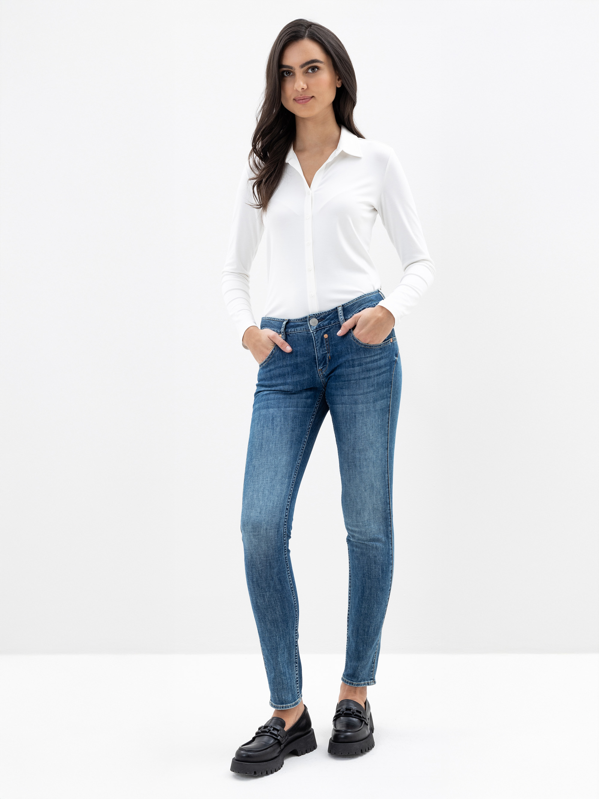 Eine Frau steht vor einem schlichten weißen Hintergrund. Sie trägt ein weißes, langärmeliges Button-up-Hemd, blaue Skinny-Jeans und schwarze Slipper. Sie hat langes dunkles Haar und ihre Hände stecken in den Taschen.