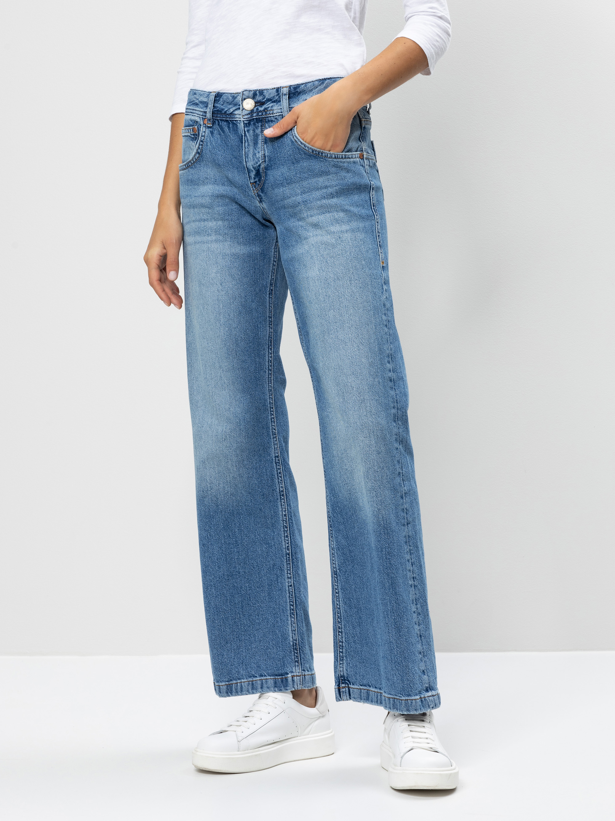 Eine Person, die hellblaue, weit geschnittene Jeans, ein weißes, langärmeliges Hemd und weiße Turnschuhe trägt, steht vor einem schlichten, hellen Hintergrund und hat eine Hand in der Tasche.