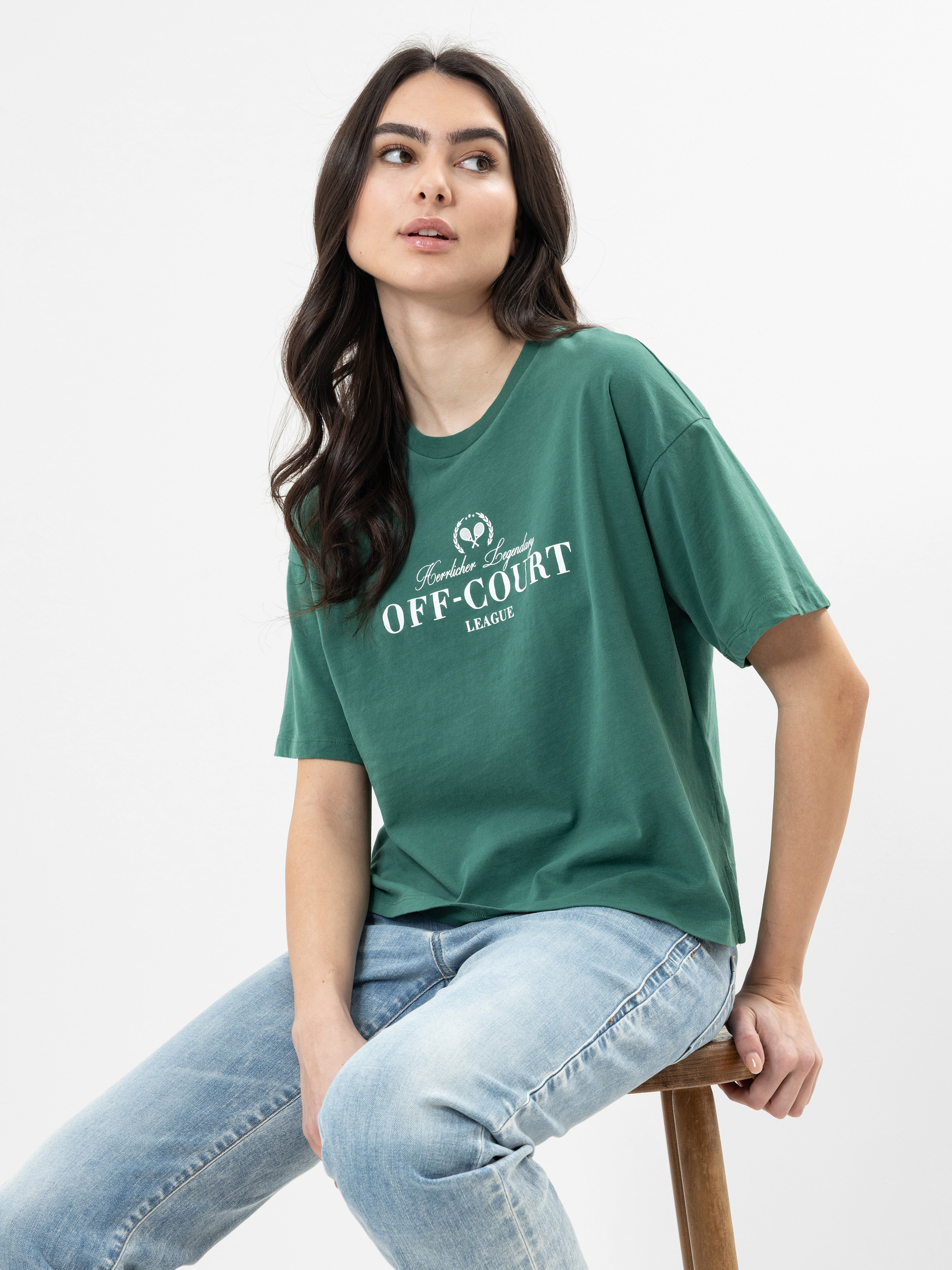 Eine Frau mit langen braunen Haaren sitzt auf einem Holzschemel und trägt ein grünes T-Shirt mit der weißen Aufschrift Heritage League OFF-COURT LEAGUE und hellblaue Jeans. Sie blickt leicht nach oben und zur Seite.