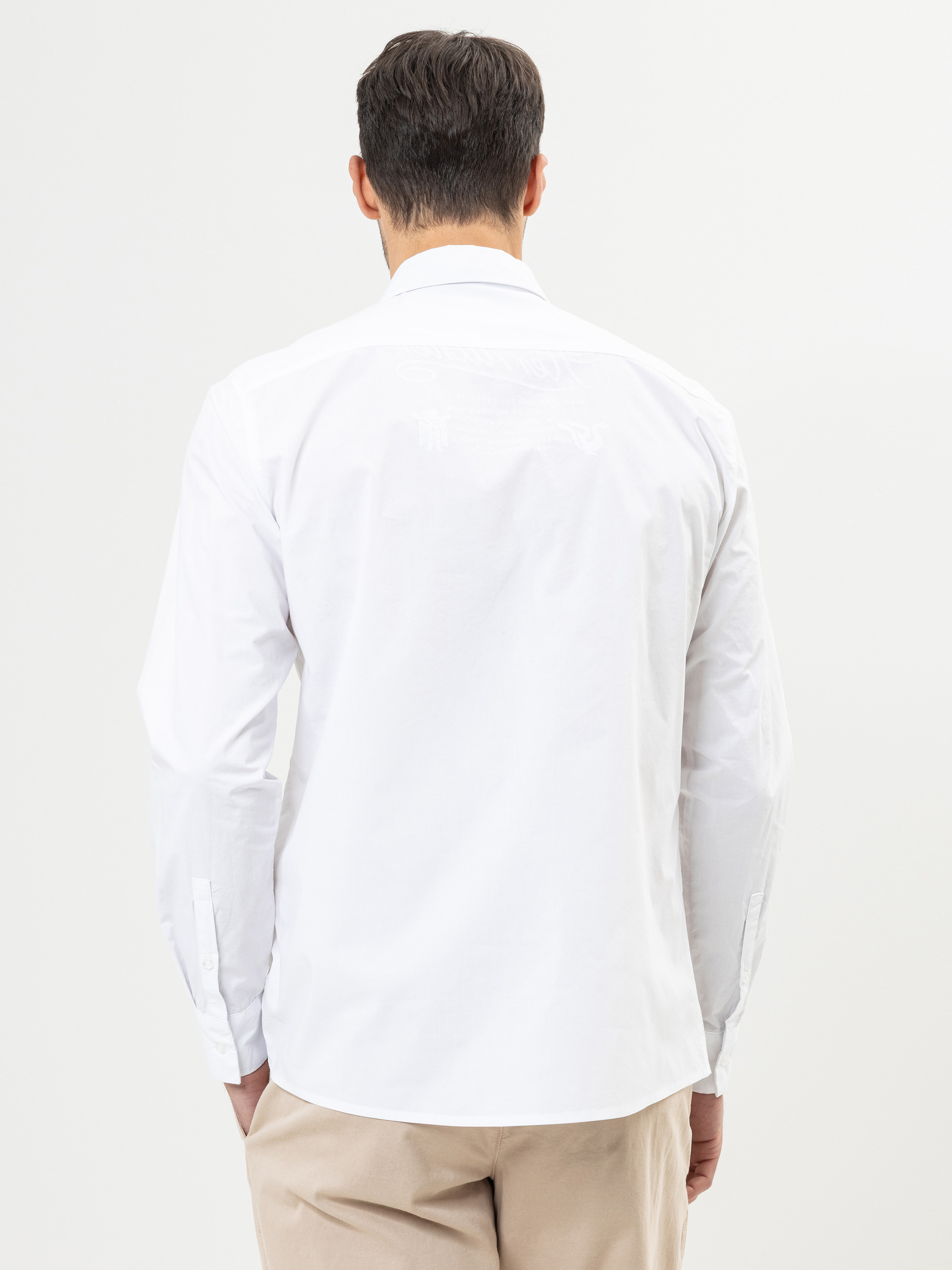 Ein Mann mit kurzen dunklen Haaren ist von hinten zu sehen. Er trägt ein weißes, langärmeliges Button-up-Hemd und eine beigefarbene Hose und steht vor einem schlichten weißen Hintergrund.